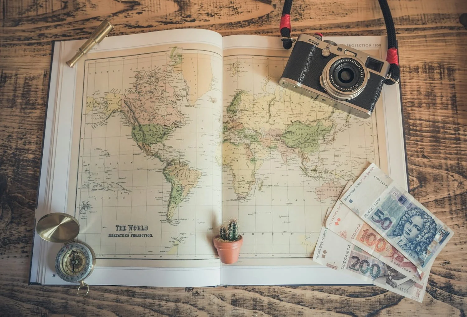 Open vintage world map with a small potted cactus in the center. On the top right, a camera with a strap. To the right, several Croatian kuna banknotes of 50, 100, and 200 denominations. On the bottom left, a compass and a gold-colored whistle. On the top left, a small silver flashlight. The items are arranged on a rustic wooden surface.