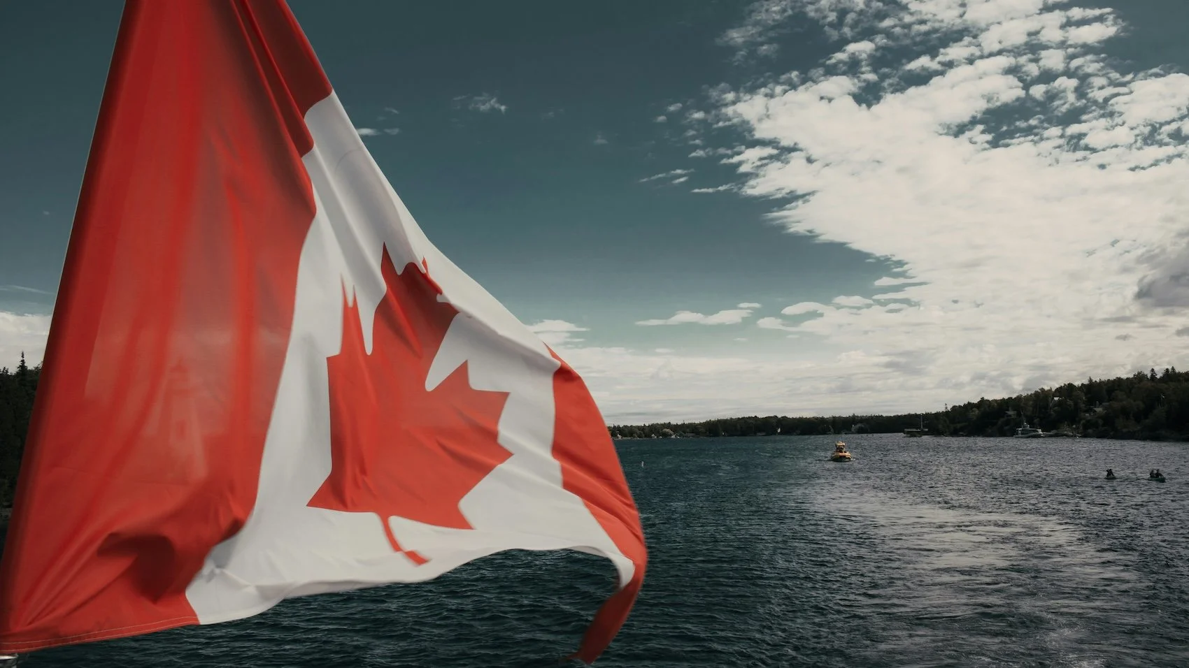 Canadian flag waving near a body of water with boats, water, and a partly cloudy sky.