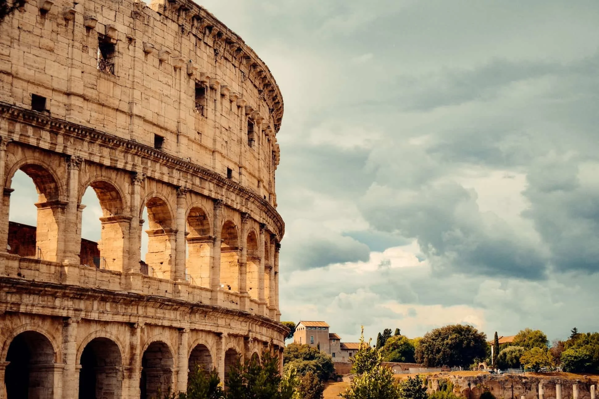 The photo shows the Colosseum in Rome, Italy, with its iconic ancient stone archways and walls. The sky is partly cloudy, and there are trees and buildings in the background.