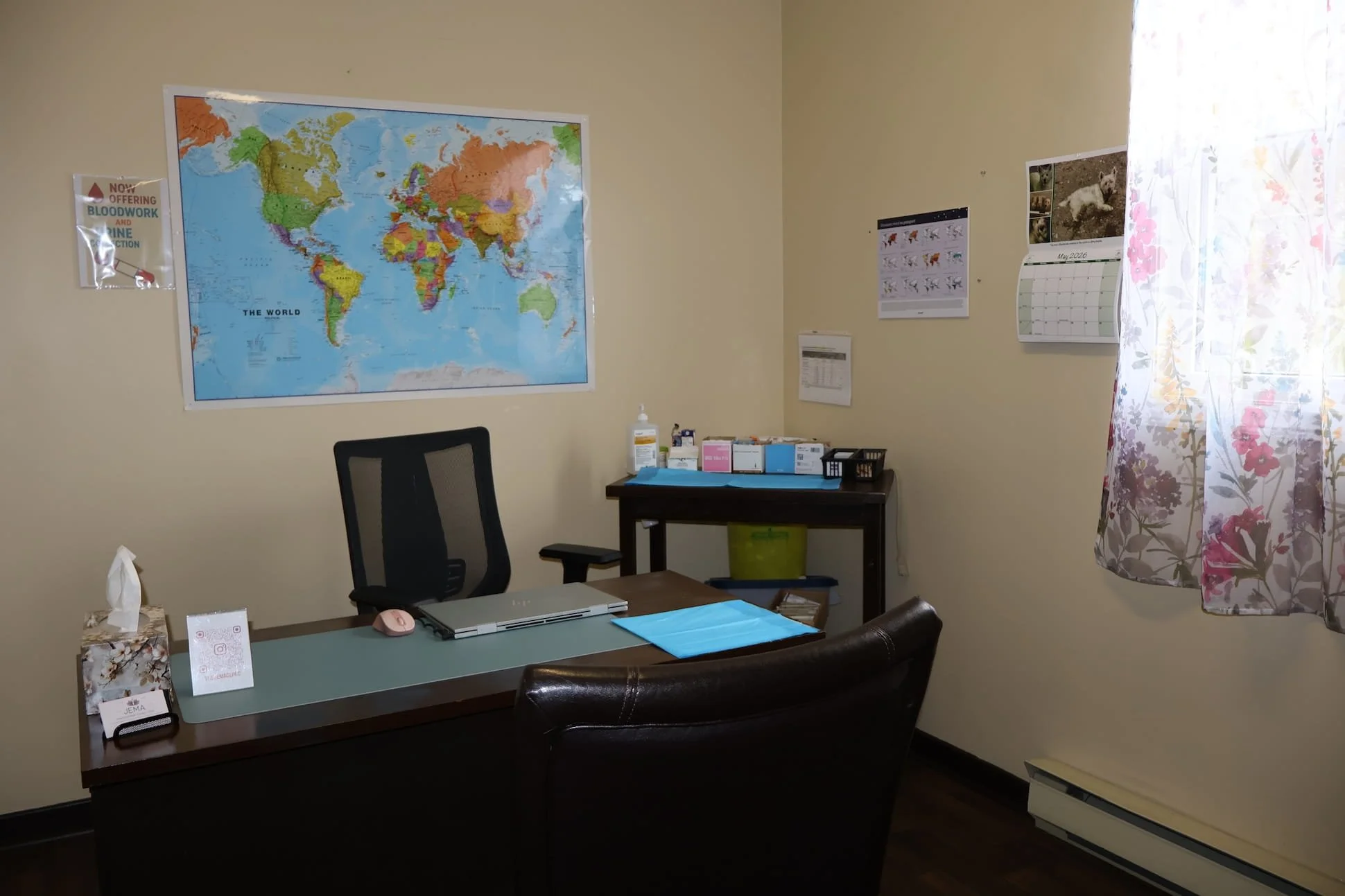 An office with a desk, chair, and a large world map on the wall. There is a calendar, hand sanitizer, and various office supplies on the wall.