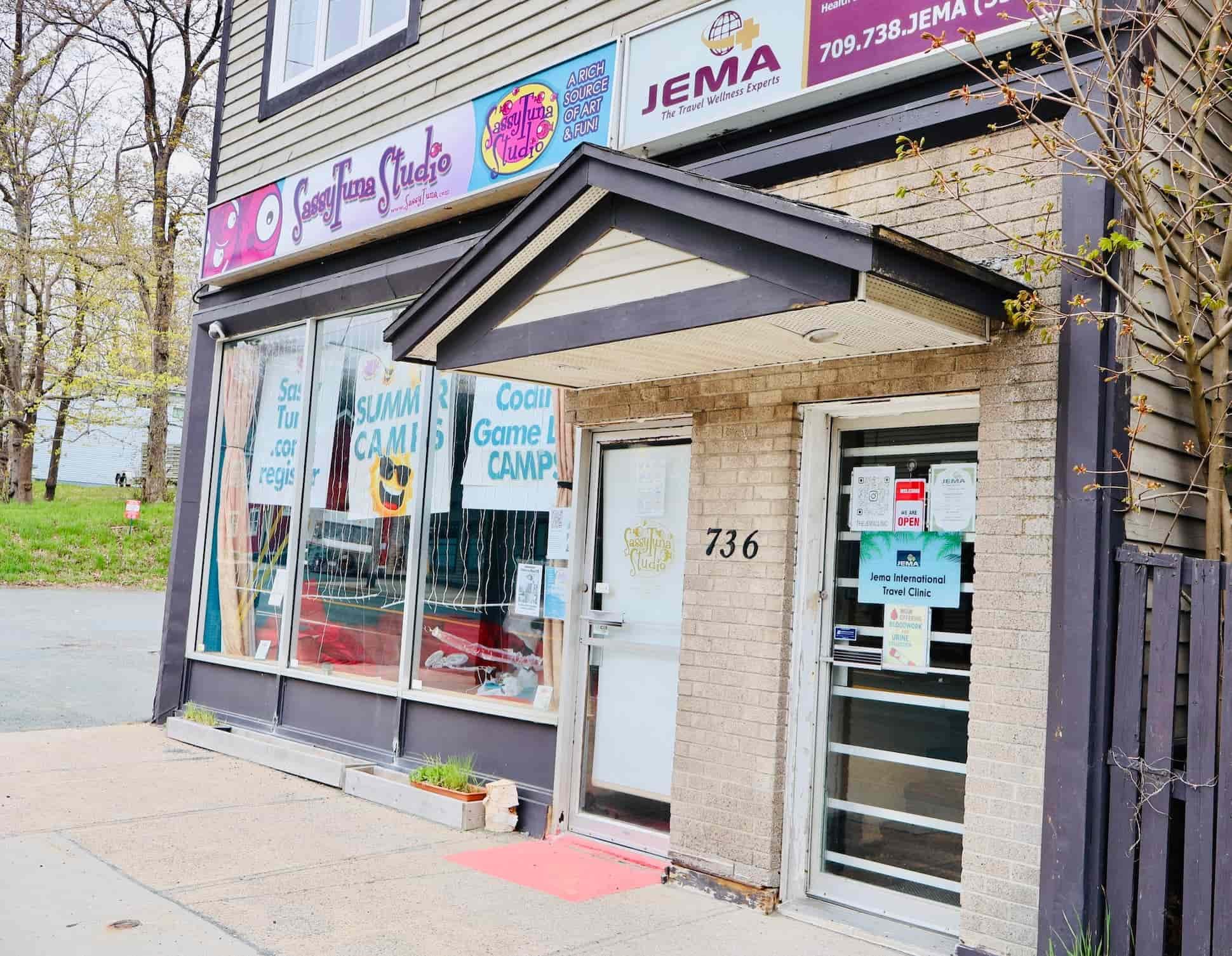 The storefront of a travel clinic named JEMA, with signage advertising art, fun, and travel wellness services. The building has a brick facade with large glass windows and a side door. There are small plants beneath the windows.
