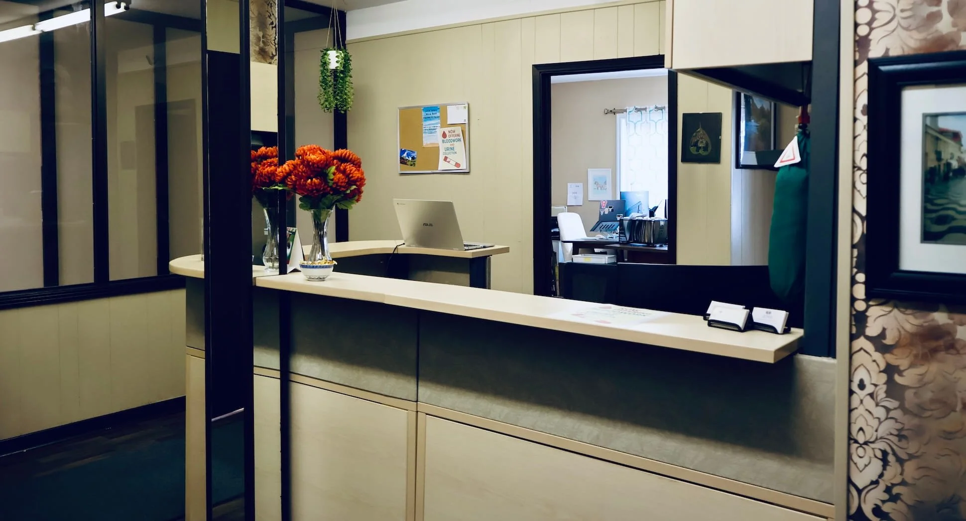 Office reception area with a curved counter, a laptop, and a vase of orange flowers, visible through a glass partition, with a doorway leading to an office with a desk and computer.