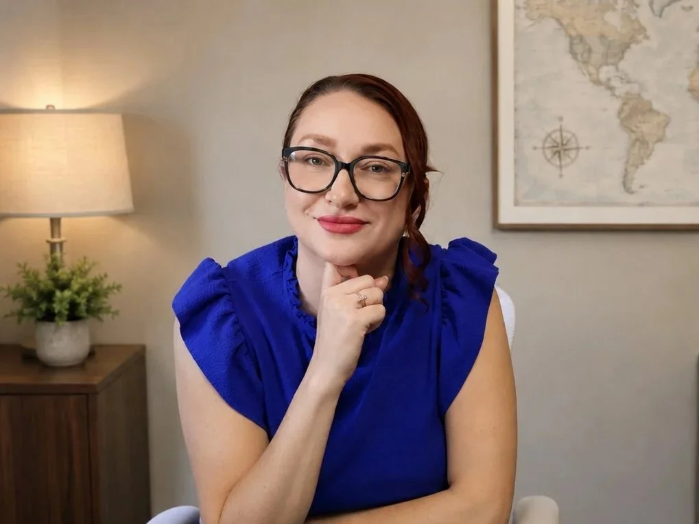 Woman with glasses and reddish-brown hair sitting in a room with a world map on the wall, a potted plant, and a lamp in the background, posing with her chin resting on her hand.