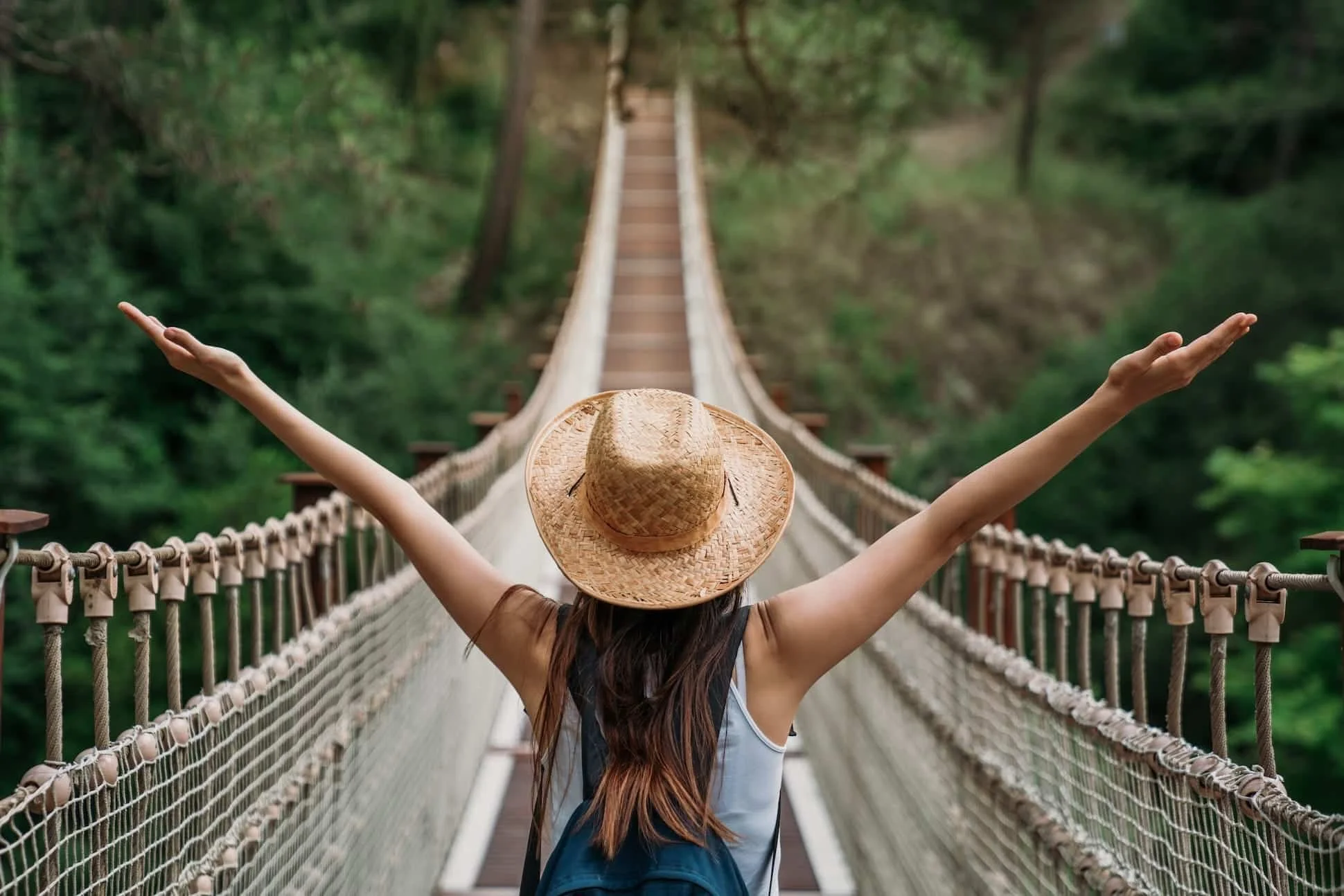 Person wearing a straw hat and a backpack crossing a suspension bridge in a lush green forest with arms raised.