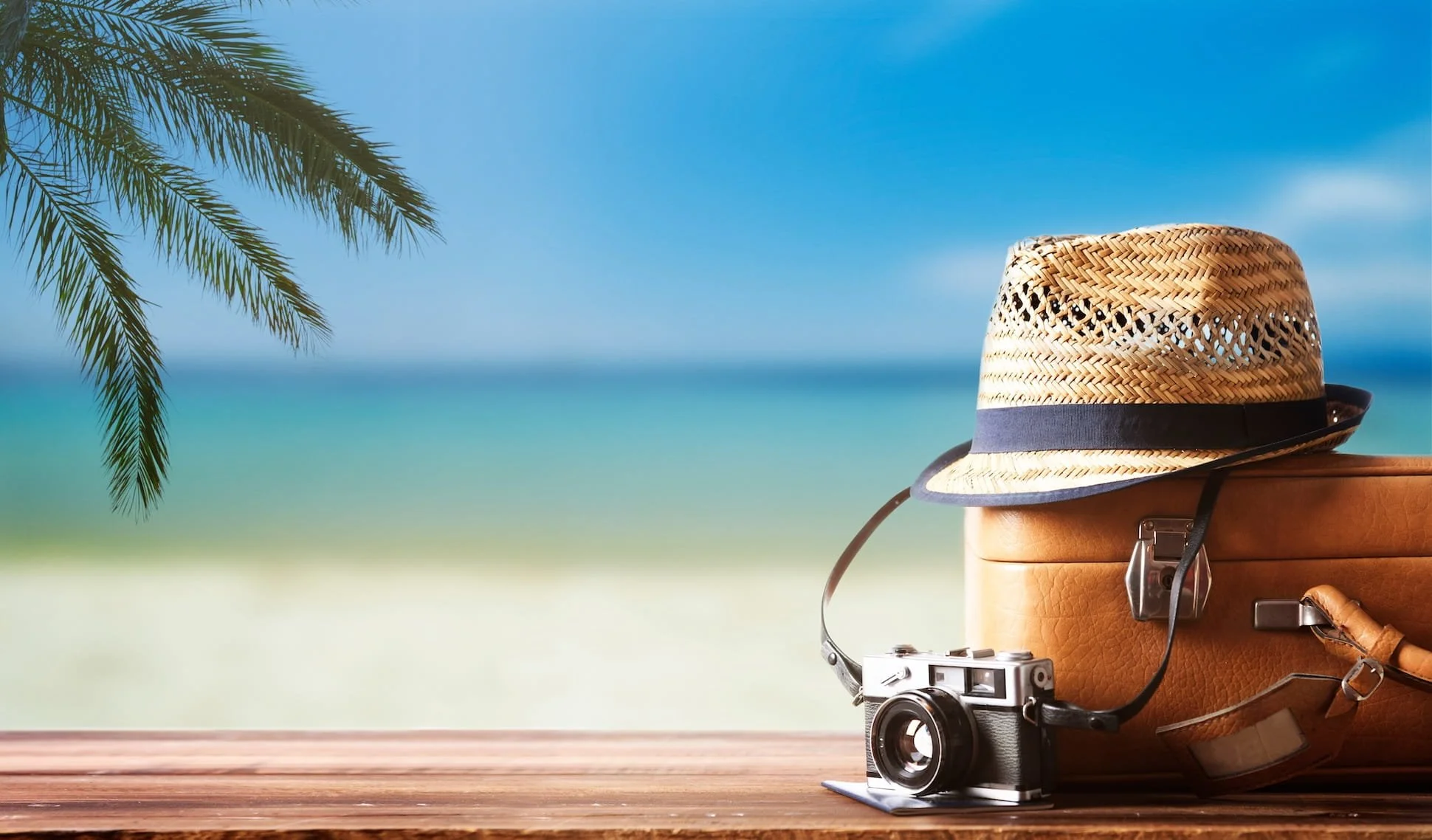 A straw hat, a vintage camera, a leather suitcase, and sunglasses are placed on a wooden surface against a beach background with a clear blue sky and palm tree branches.
