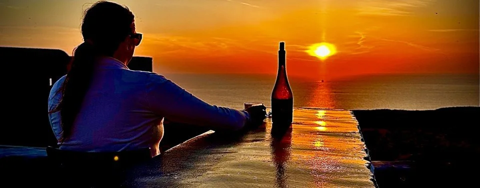 A woman with a ponytail and glasses sitting at a table overlooking the sunset over the ocean, with a bottle of wine and a glass in front of her.