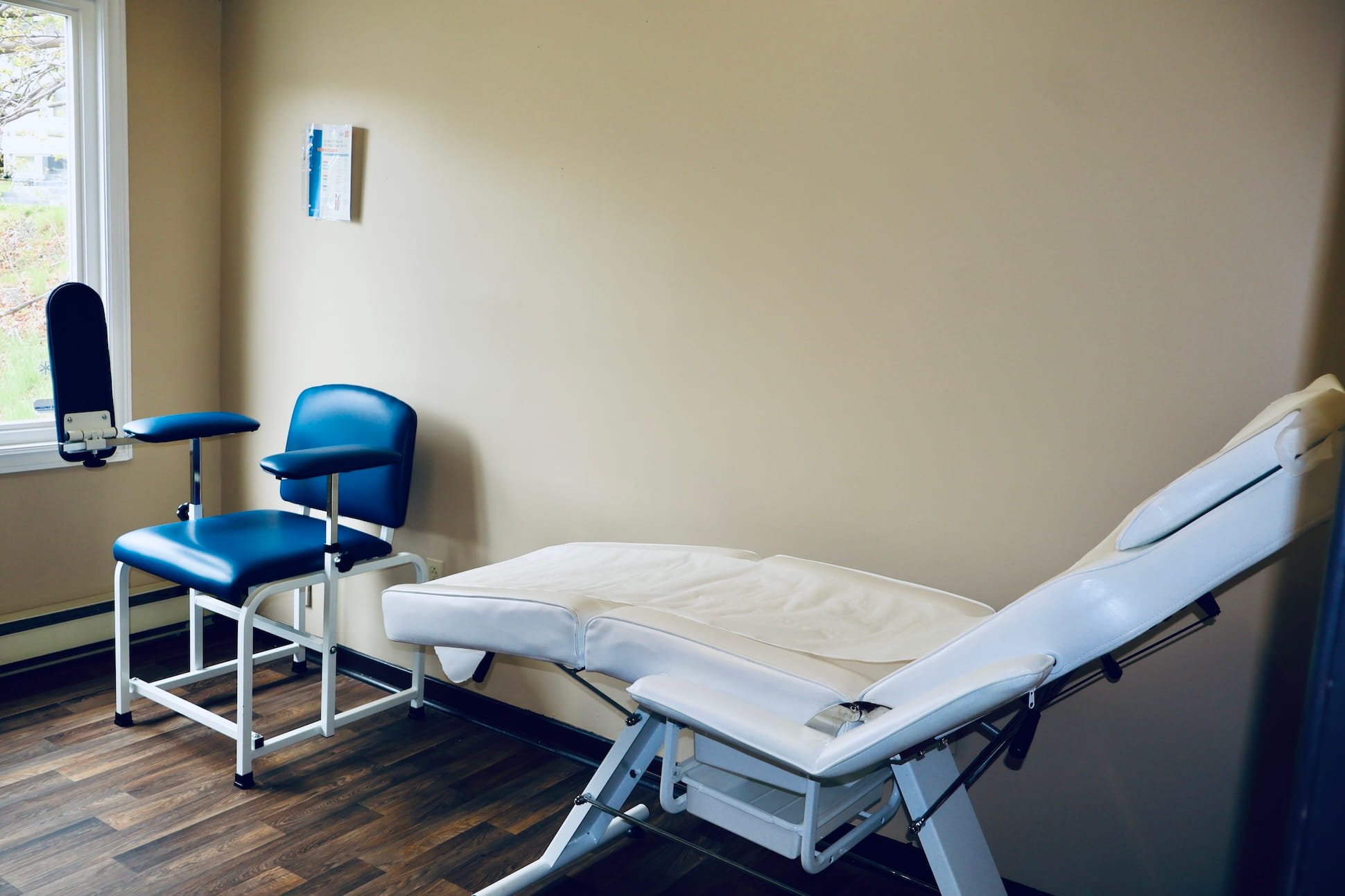 A hospital room with a bed, a blue chair, and a patient lift chair near the bed. There is a window letting in natural light and a wall-mounted chart or information sheet.