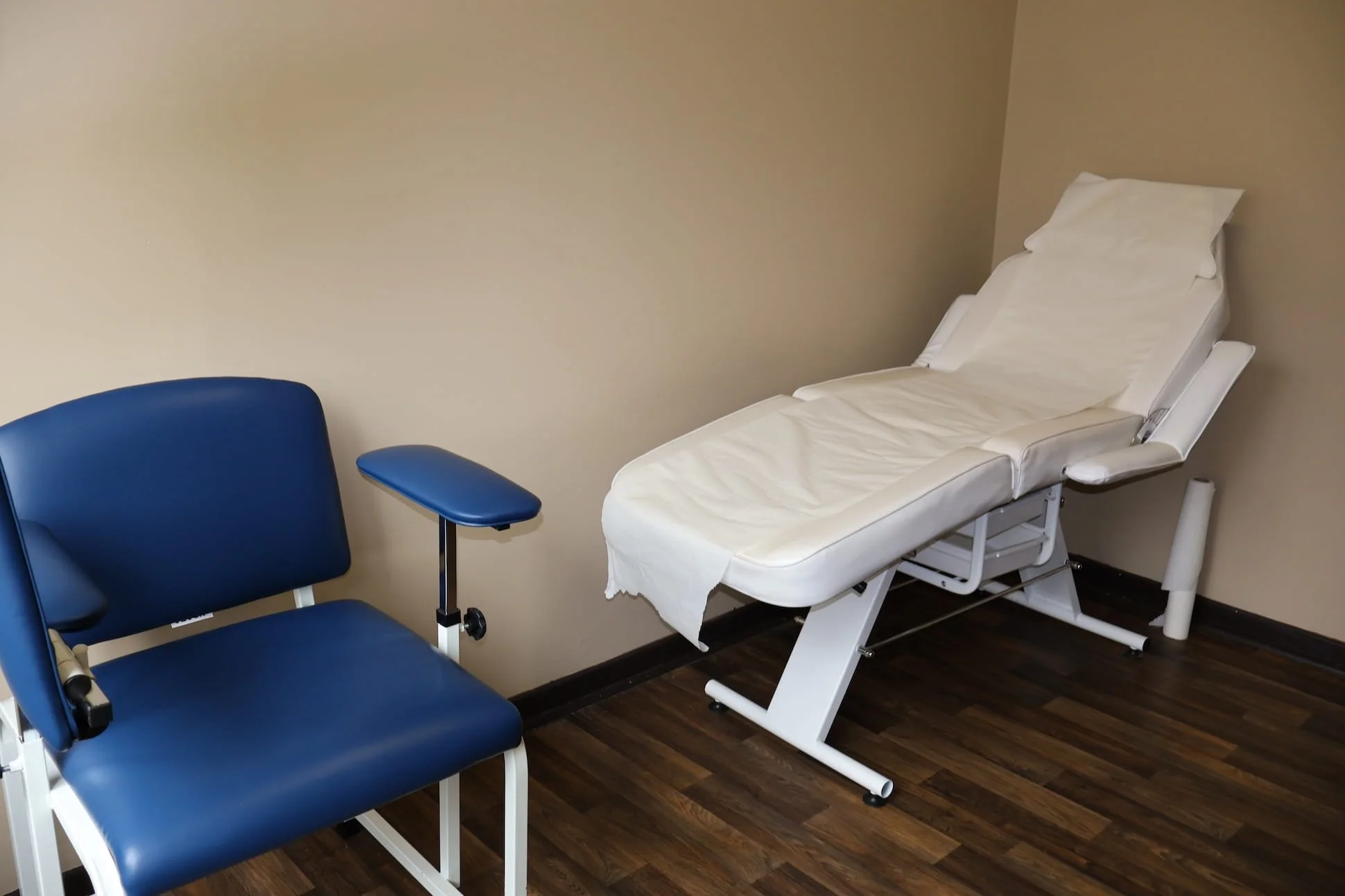 Medical examination room with a blue chair, adjustable exam table with white paper sheet and pillow, and beige walls.