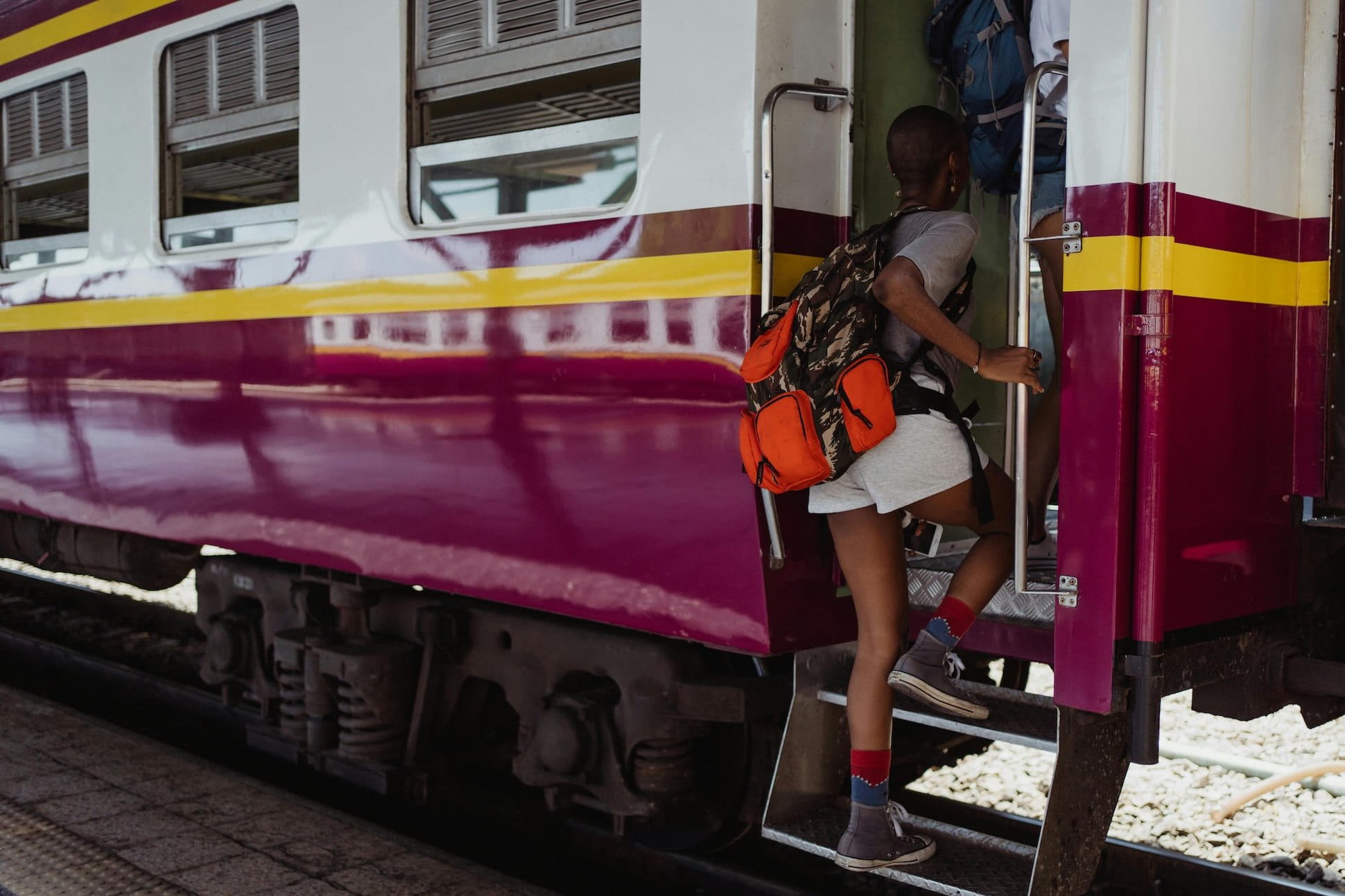 A person is boarding a maroon and yellow train, climbing the steps with a backpack, wearing gray and red socks, gray shorts, and a gray t-shirt.