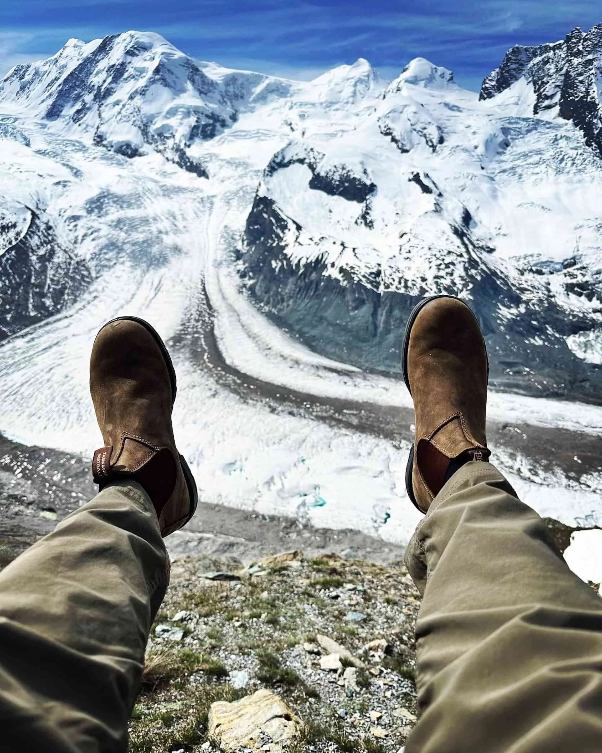 Person sitting on rocky ground with snow-capped mountains and glaciers in the background, wearing brown boots and beige pants.