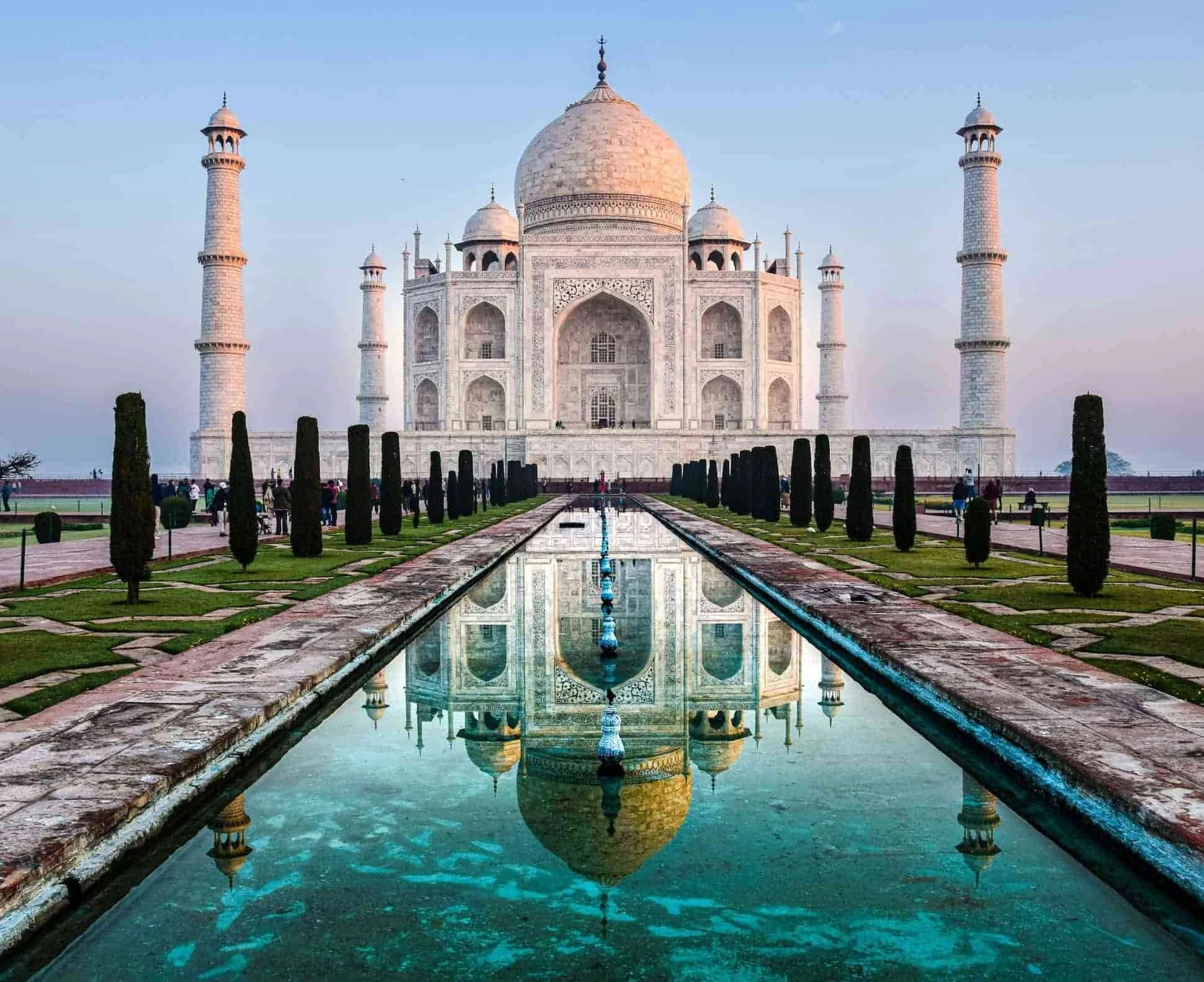 The Taj Mahal at dawn with a reflection in the water pool in front, surrounded by greenery and trees.