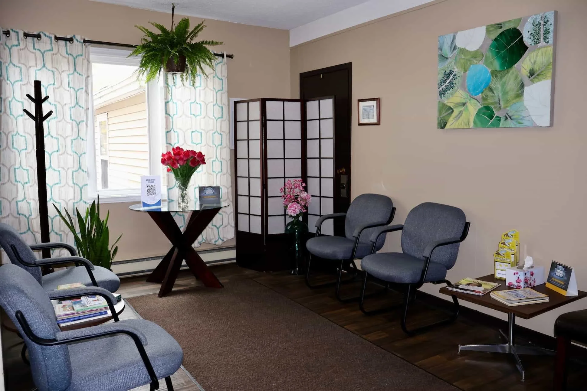 A waiting room with gray chairs, a small wooden table with magazines, a round glass table with a bouquet of red roses and tropical leaves, a plant, a folding room divider, and wall art with green leaves.