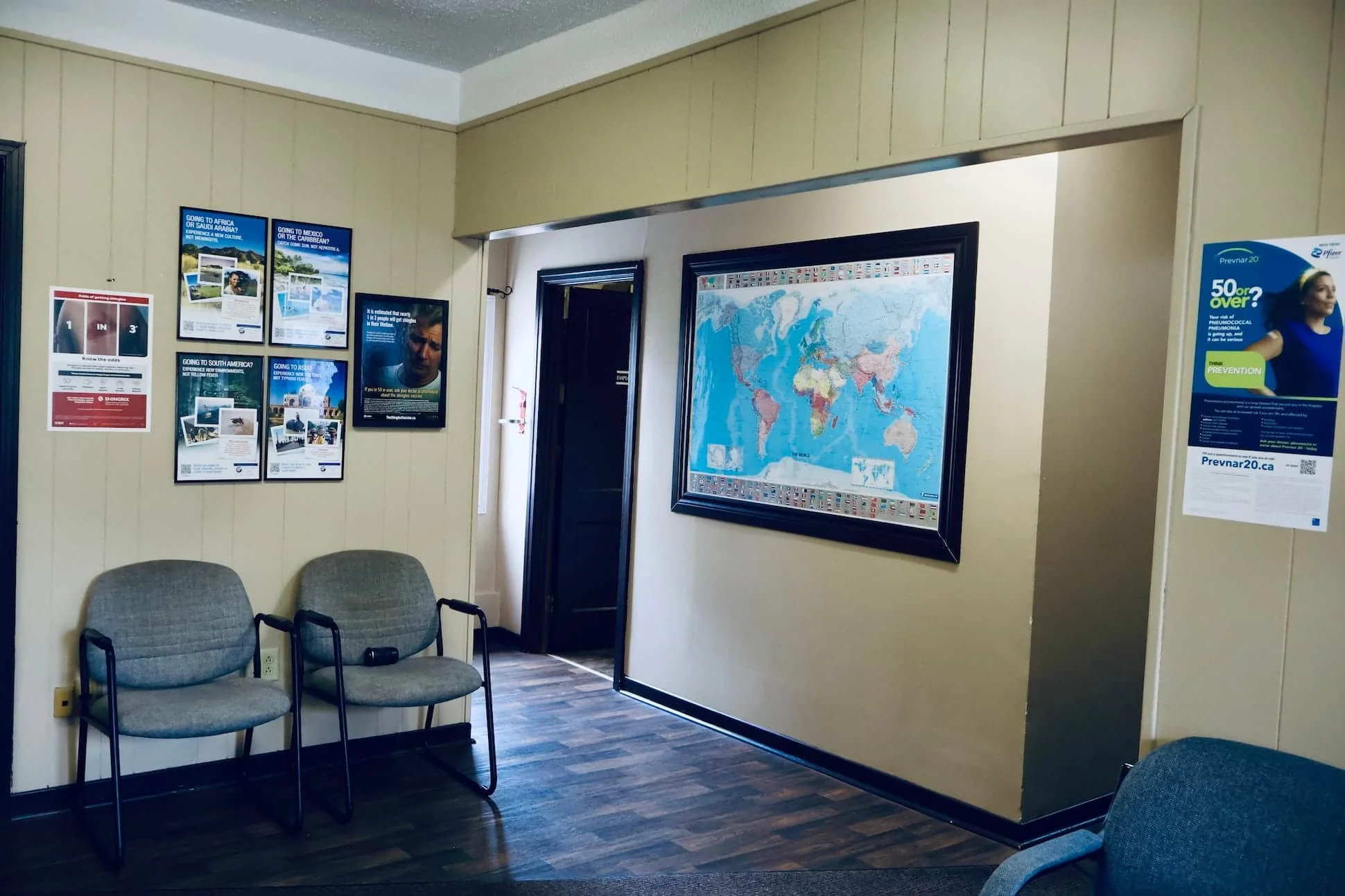 Waiting room with two gray chairs, wall posters about travel, a world map, and a door leading to another room.