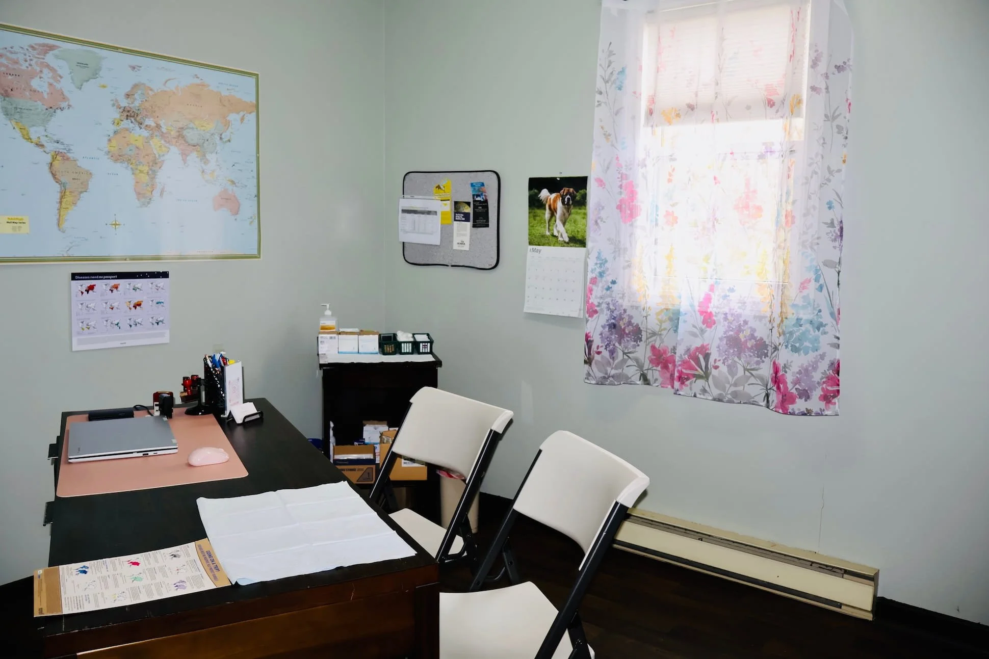 An office with a black desk, white chairs, a map on the wall, a calendar, and a window with floral curtains.