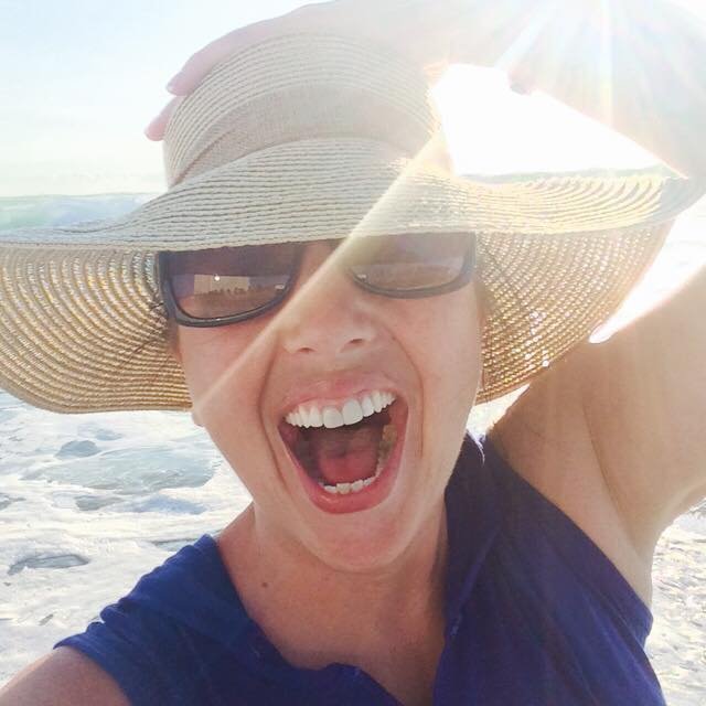 Woman at the beach wearing sunglasses and a wide-brimmed hat, laughing with her mouth open, with ocean waves in the background and sunlight shining through.