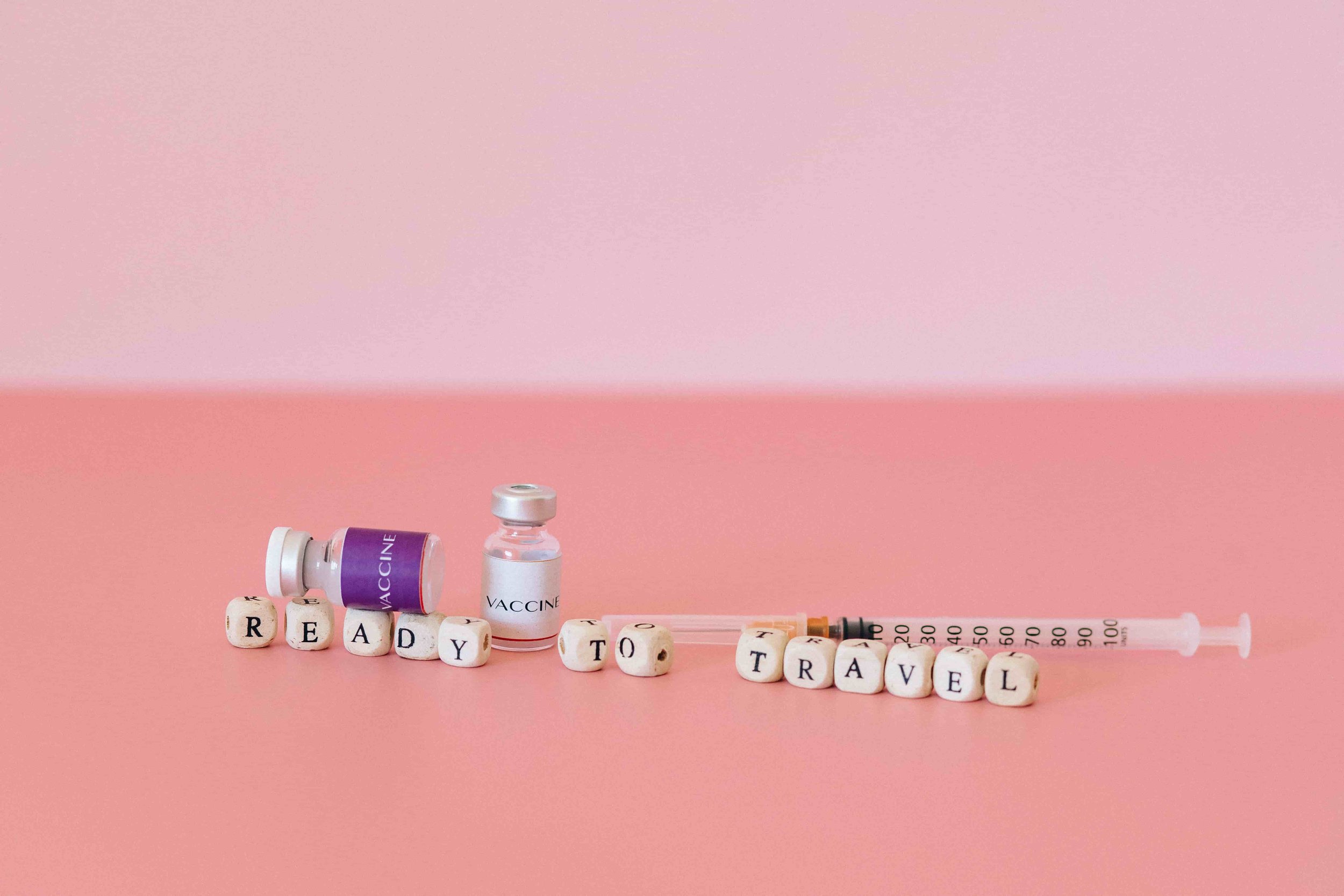 Vaccine vials, a syringe, and lettered blocks spelling out 'READY TO TRAVEL' on a pink background.