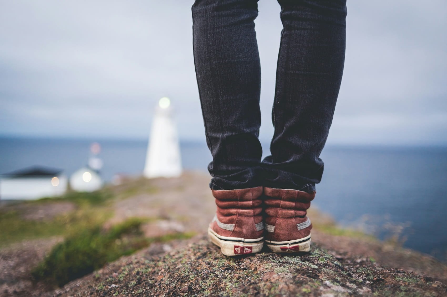 Close-up of a person wearing worn shoes standing on a rocky surface near a body of water, with a lighthouse in the background on a cloudy day.