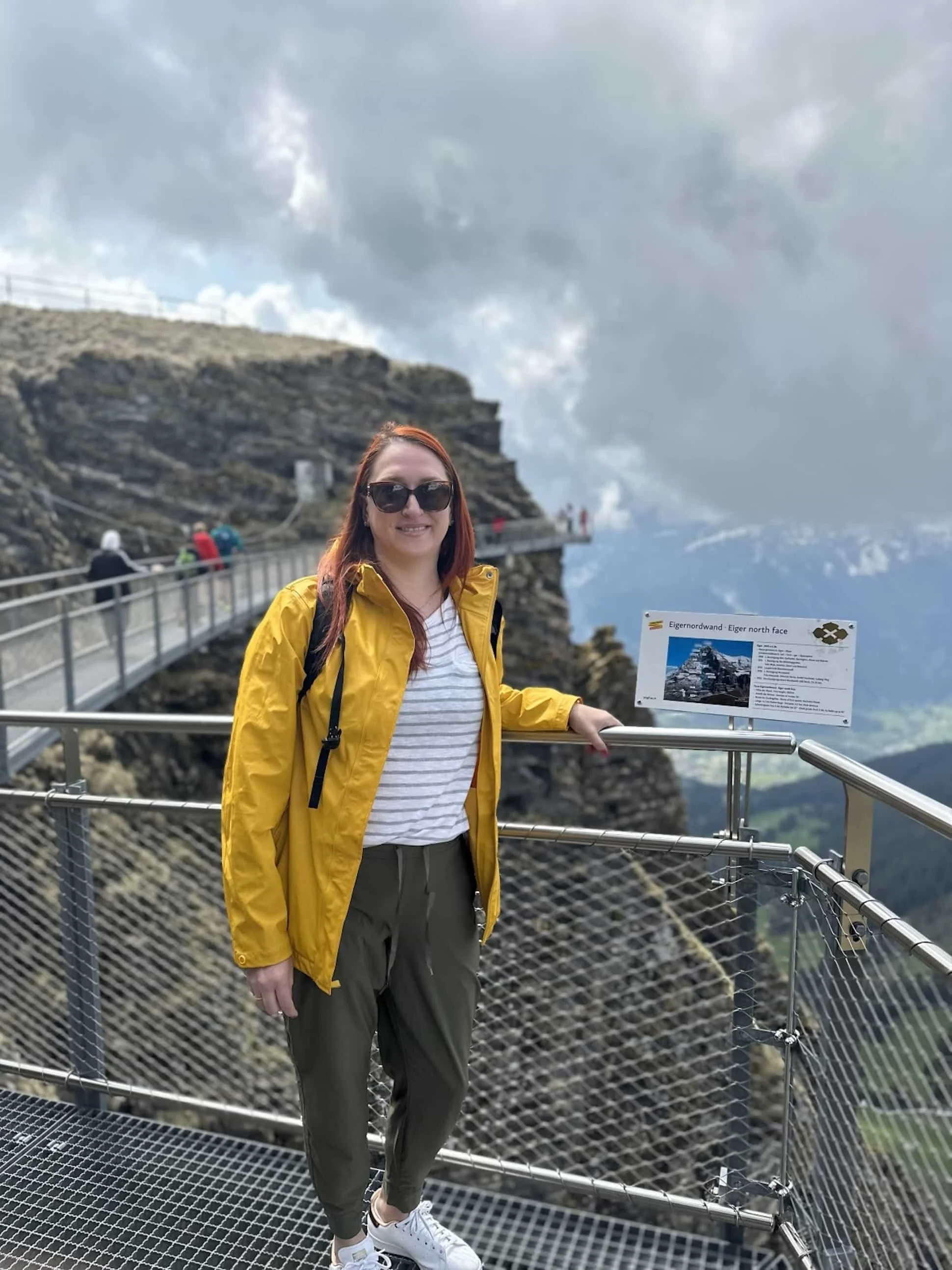 A woman with red hair wearing sunglasses, a yellow jacket, a white striped shirt, and green pants, standing on a metal observation platform at Eiger, North Face, with cloudy mountain scenery in the background.