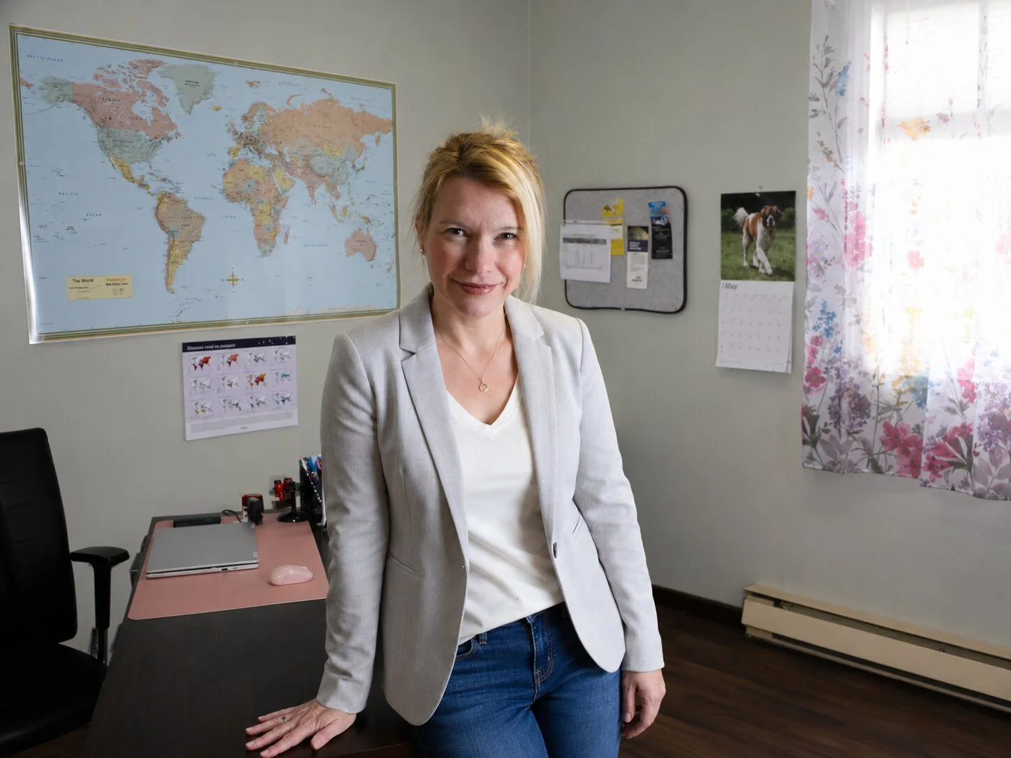 A woman standing in an office, wearing a light gray blazer and white top, smiling at the camera. Behind her on the wall is a world map, calendar with a dog photo, corkboard with papers, and a window with floral curtains.