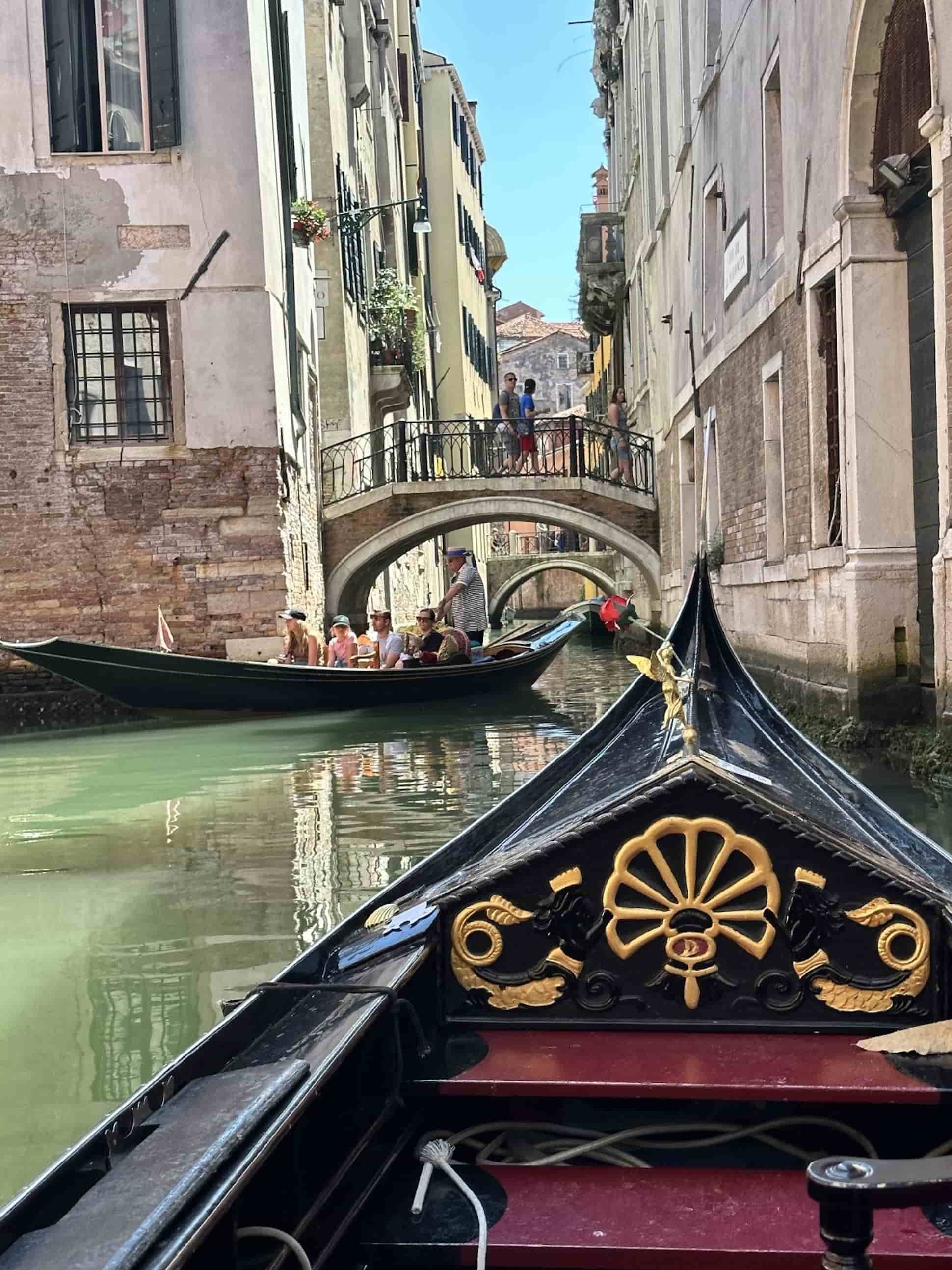 View from a gondola navigating through a small canal in Venice, Italy, with tourists on another gondola and people walking across a small bridge between historic buildings.