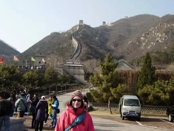 A woman in a red jacket and sunglasses standing in front of the Great Wall of China, with tourists and cars nearby, surrounded by trees and hills.