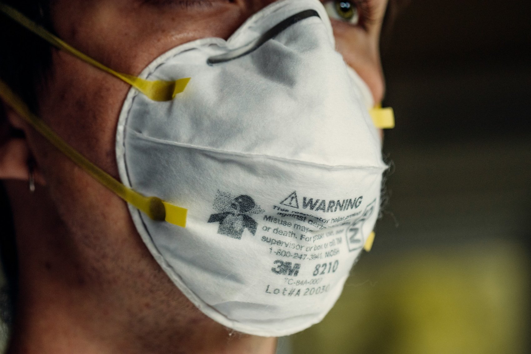 Close-up of a person wearing an N95 mask with yellow straps, partially covering their face, showing one eye and some facial skin.