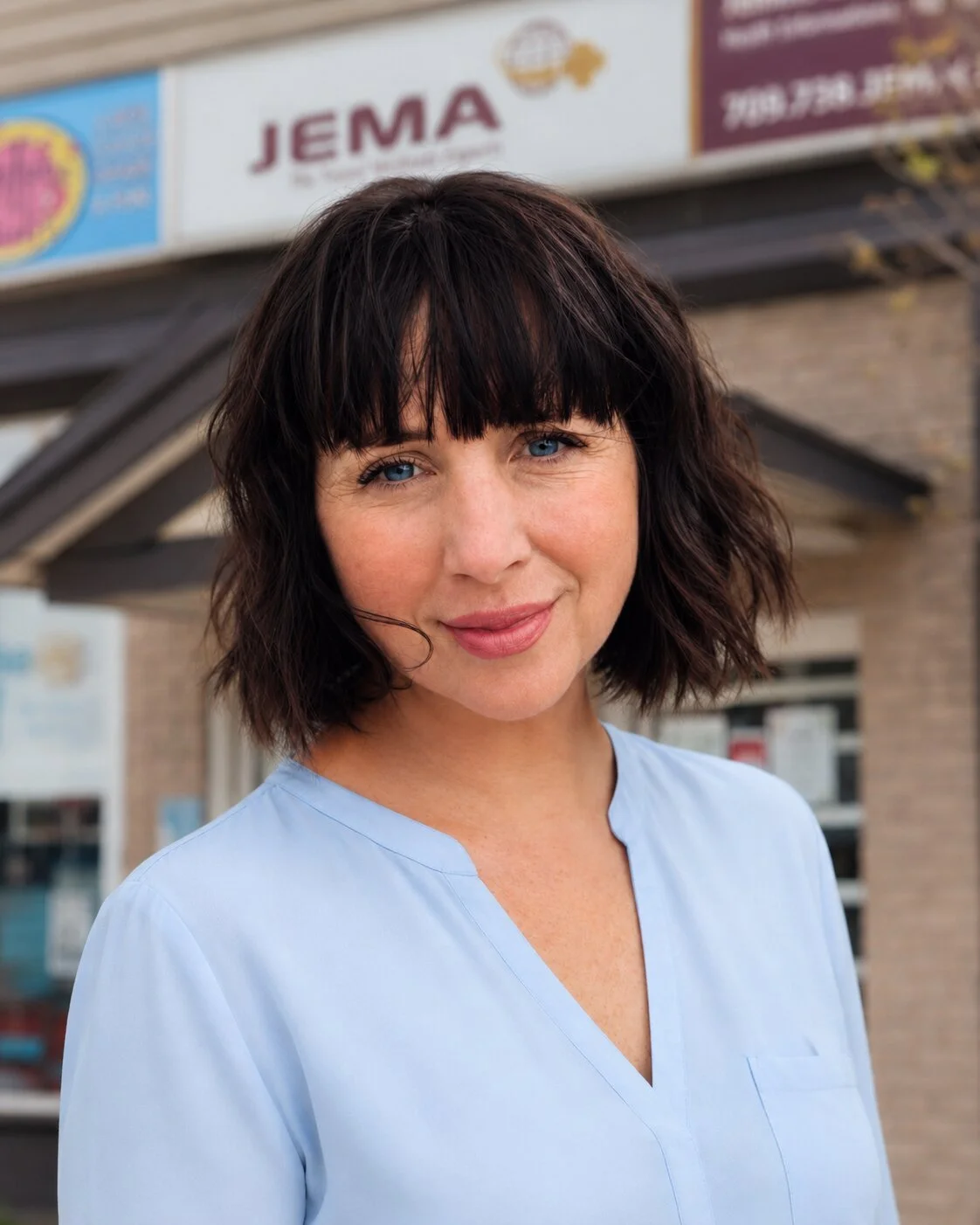 A woman with shoulder-length dark wavy hair with bangs, blue eyes, and a light blue medical uniform standing outdoors in front of a building with signs, one of which says 'JEMA'.
