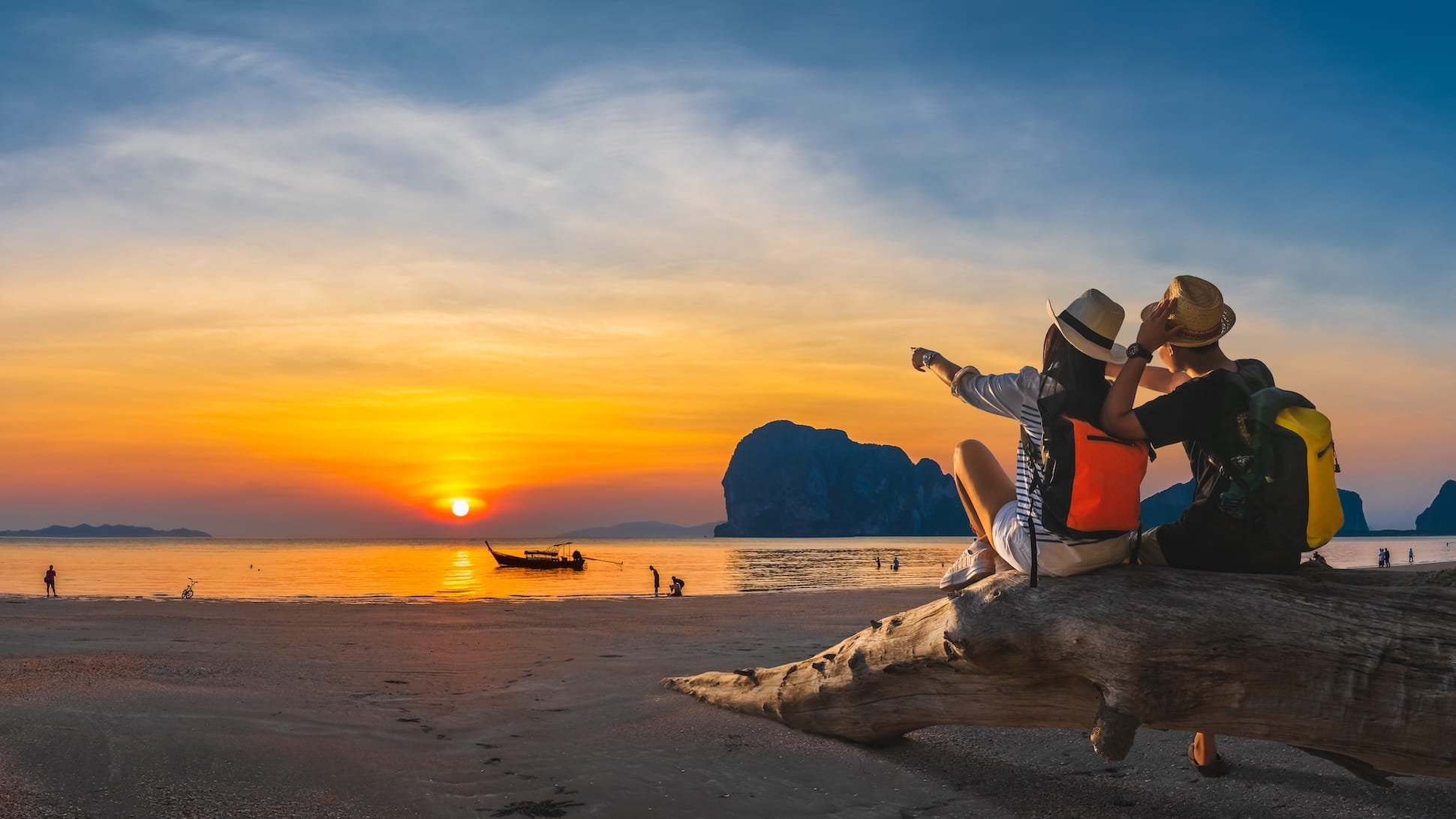 Two people sitting on a large piece of driftwood on a beach during sunset, pointing towards the horizon. They are wearing hats and backpacks, with a boat and a few people visible in the water and on the shore.