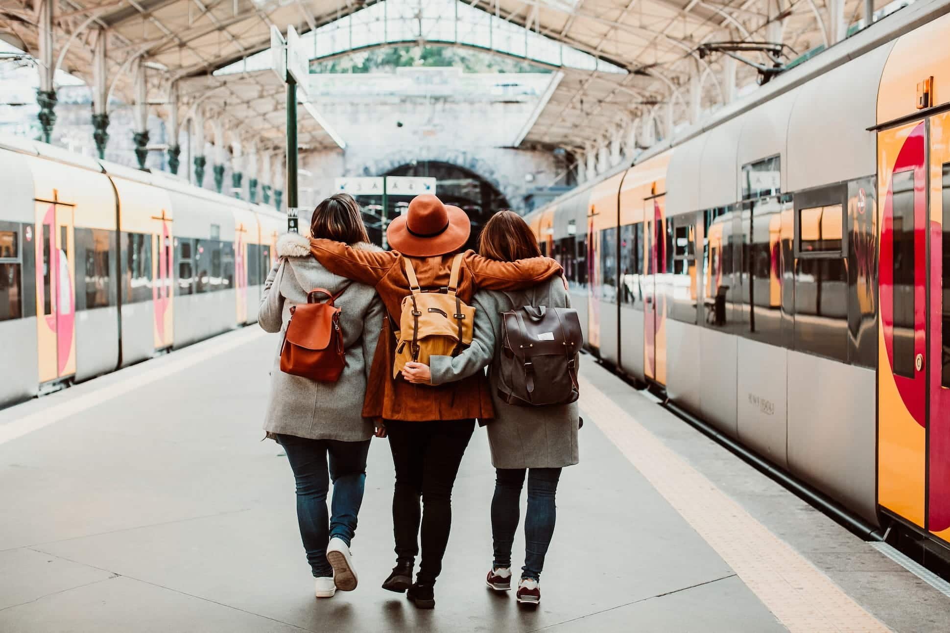 Three women are walking together at a train station, with their backs facing the camera, arms around each other. They are in front of a yellow and silver train, and the station has an arched roof with steel beams.