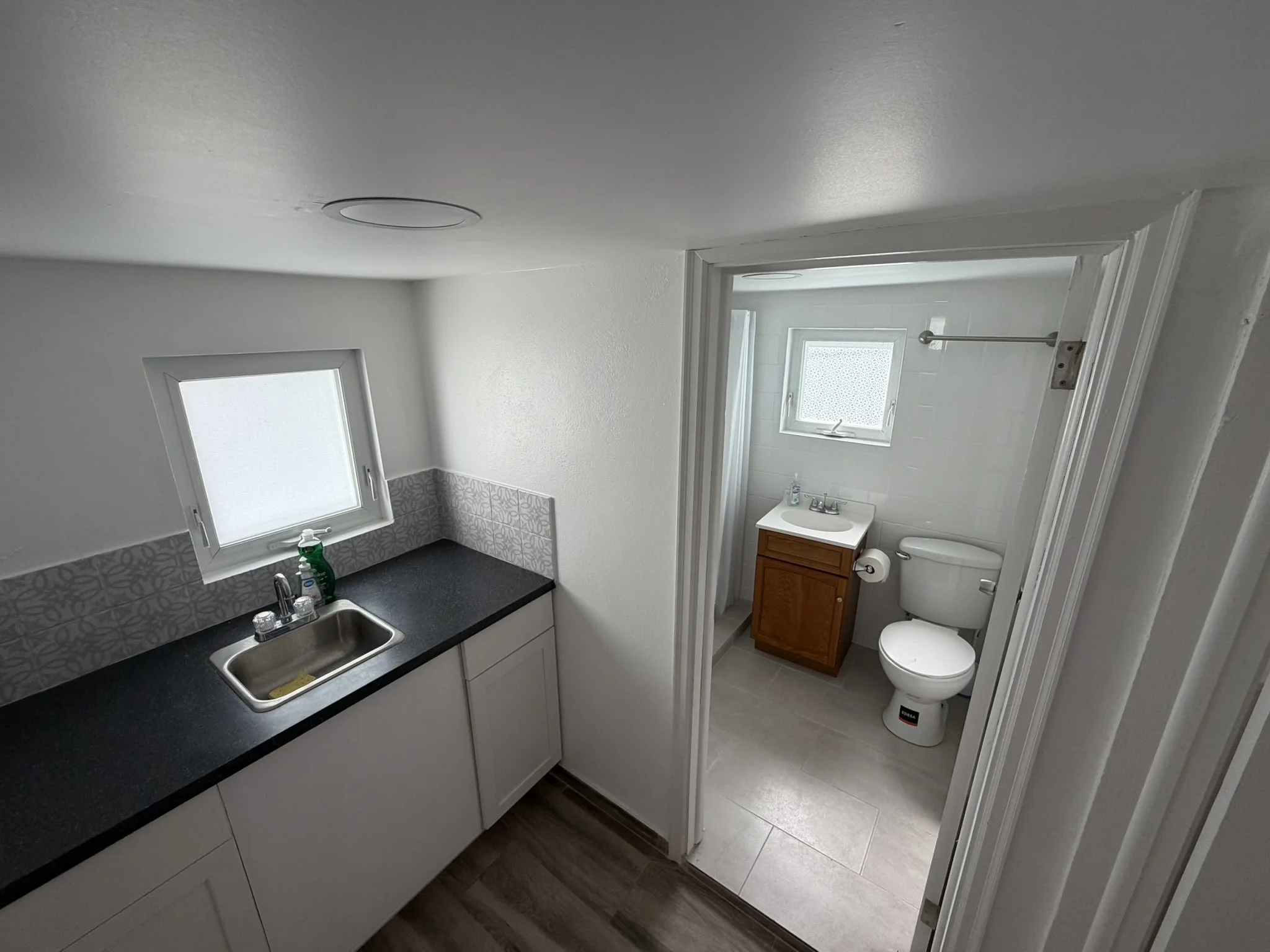 A small kitchenette with a black countertop, white cabinets, and a window, adjacent to a bathroom with a small sink, toilet, and another window.