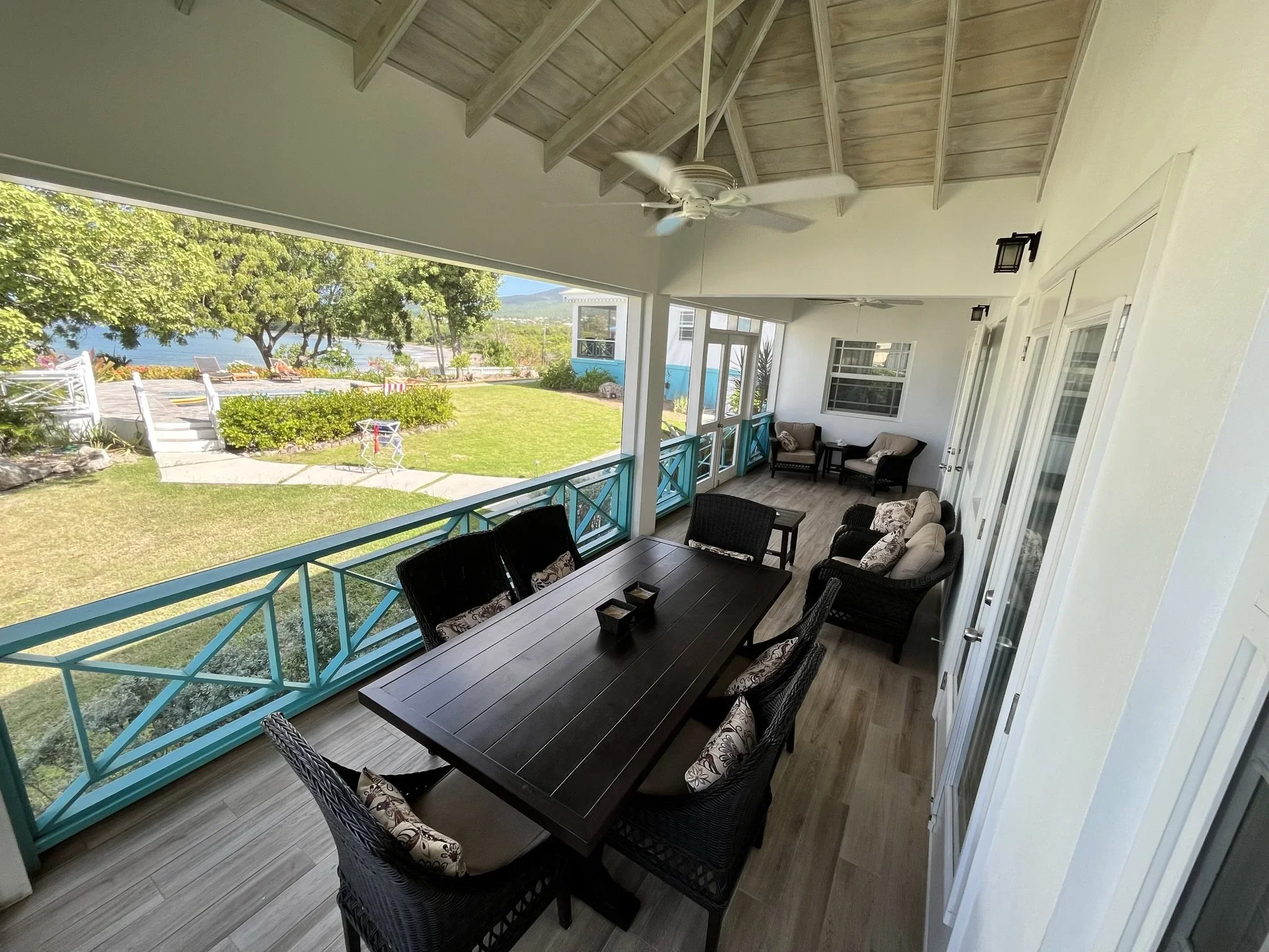 Covered porch with a dining table and eight chairs, a sitting area with two cushioned chairs and a small table, ceiling fans, and a view of the lawn, pool and the Caribean sea in the background.