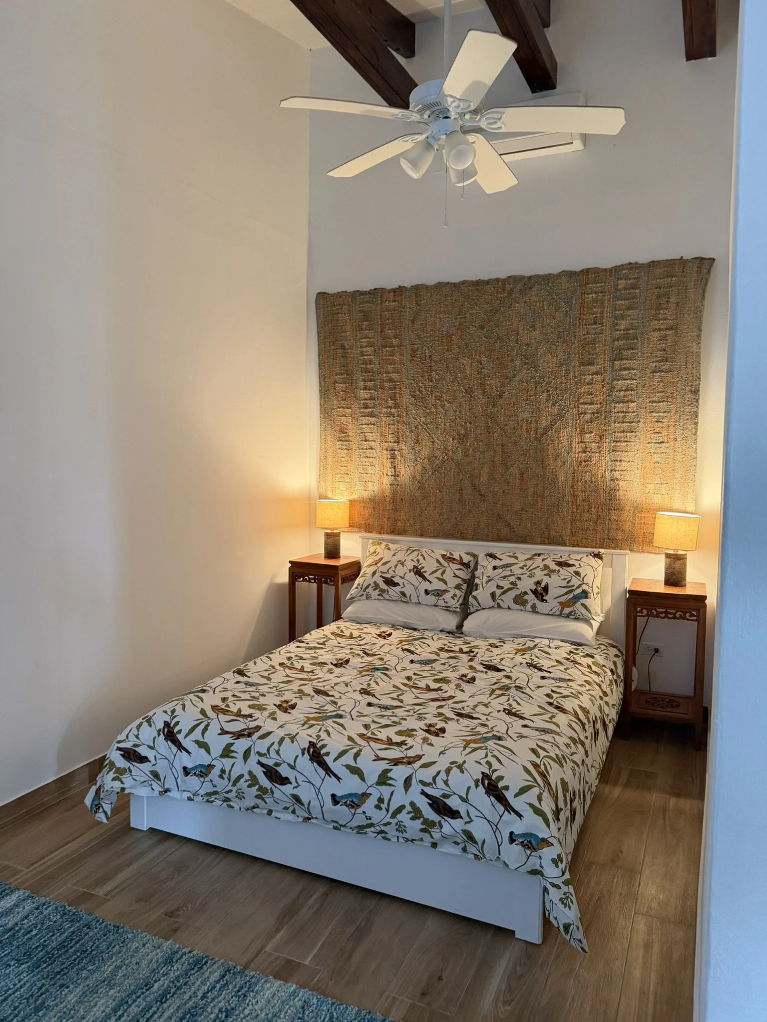 A cozy bedroom with a bed featuring a bird-patterned duvet and pillows, two wooden nightstands with lamps, a textured headboard, a ceiling fan, and wooden flooring.