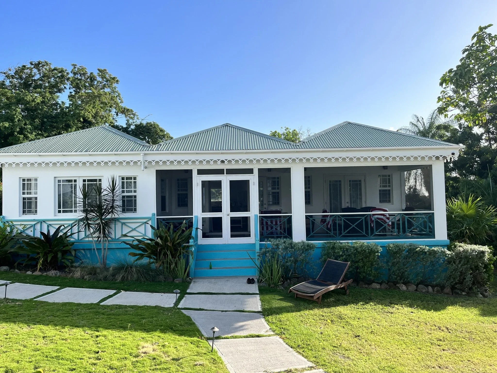 Beautiful Caribean cottage with a white exterior, light blue trim, and a metal roof, featuring a front porch with chairs, surrounded by green plants and a well-maintained lawn.