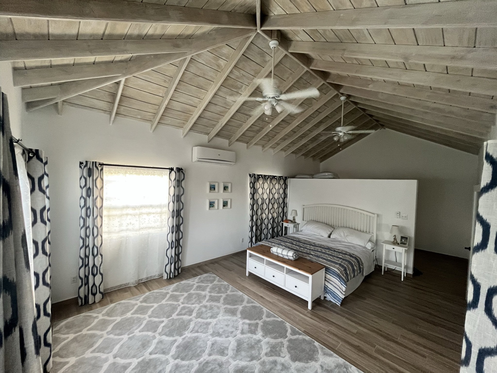 A spacious bedroom with a vaulted wooden ceiling, two ceiling fans, and light-colored walls. It features a large bed with white headboard, striped bedding, and a bench at the foot of the bed. There are nightstands with lamps on each side of the bed, a white area rug on the wooden floor, a window with patterned curtains, and four small framed pictures on the wall above the bed.