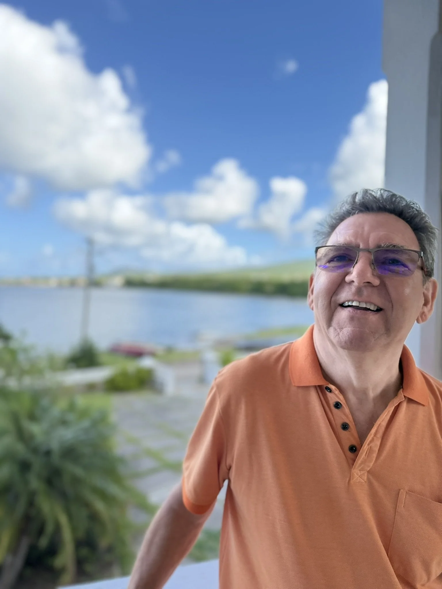 John wearing glasses and an orange polo shirt standing outdoors on a balcony, smiling with a view of a the Caribean sea, blue sky, and white clouds in the background.