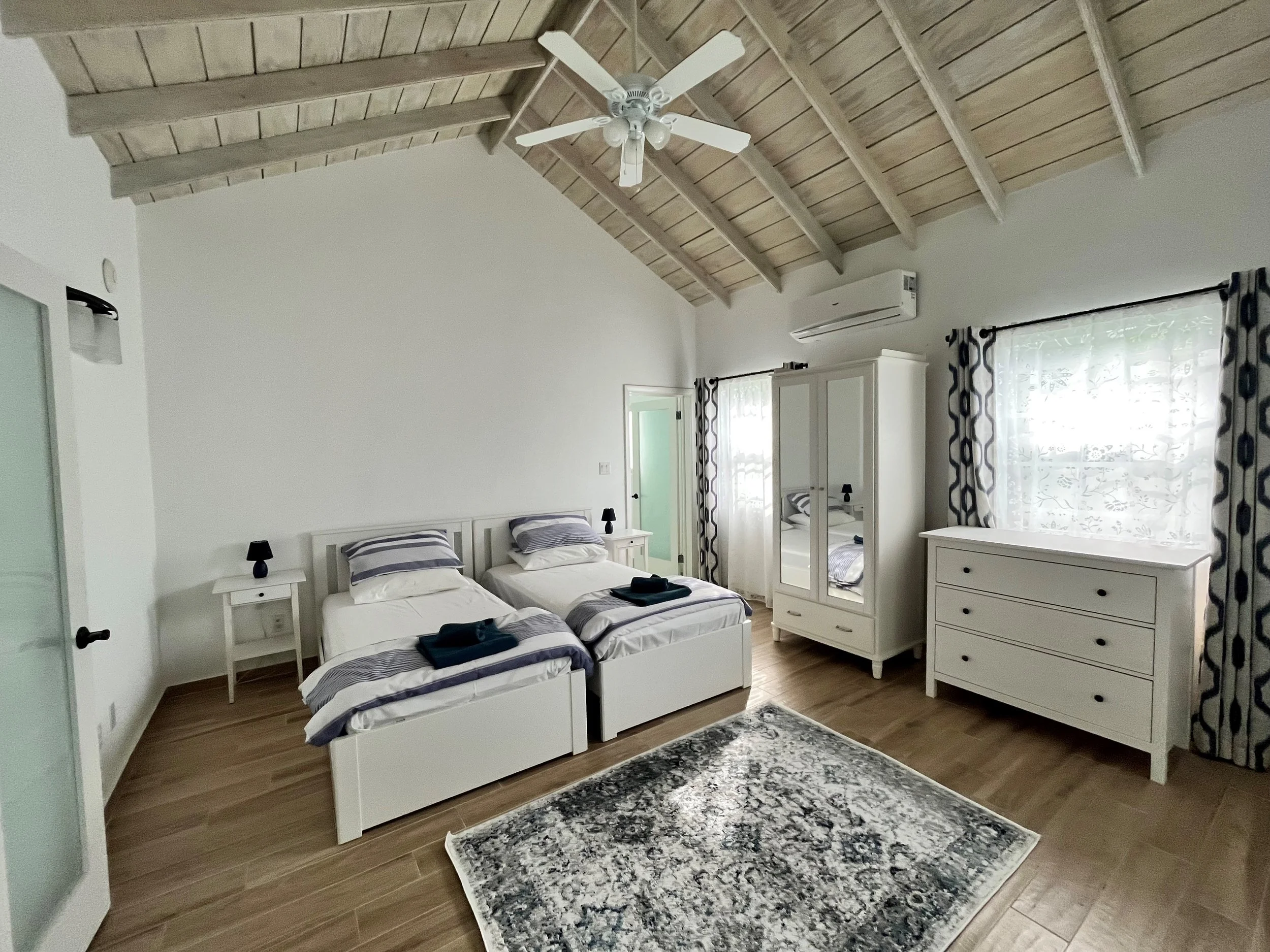 A bedroom with two twin beds, a white wardrobe with mirrored doors, a white dresser, an air conditioning unit, and windows with patterned curtains. The ceiling has wood paneling and a white ceiling fan, and the floor is made of hardwood tiles.