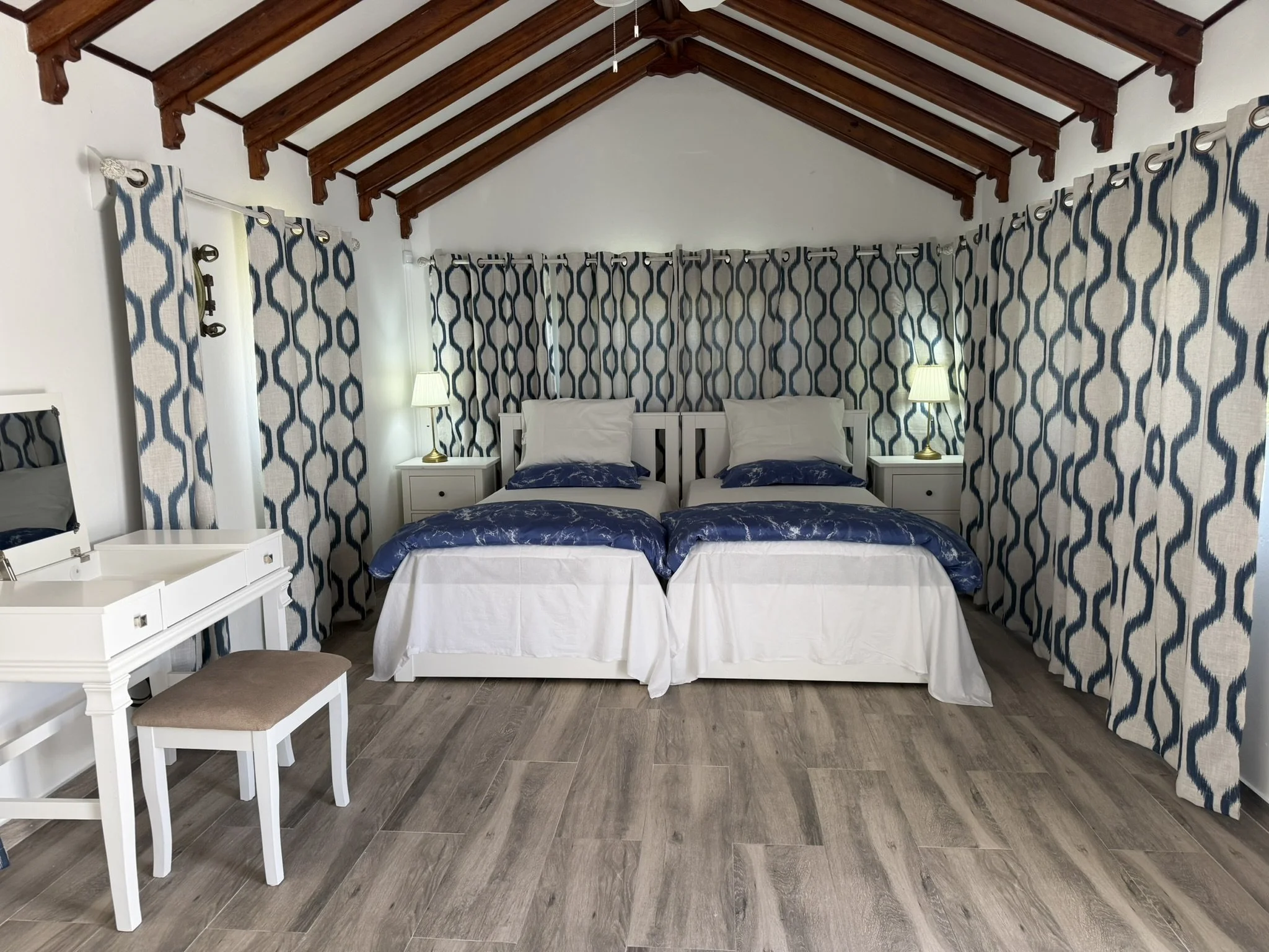 Stunning bedroom with two twin beds, blue patterned comforters, and white headboards, flanked by nightstands with lamps. Blue and white patterned curtains cover windows beneath a wooden-beam ceiling, with a white vanity and stool in the foreground.