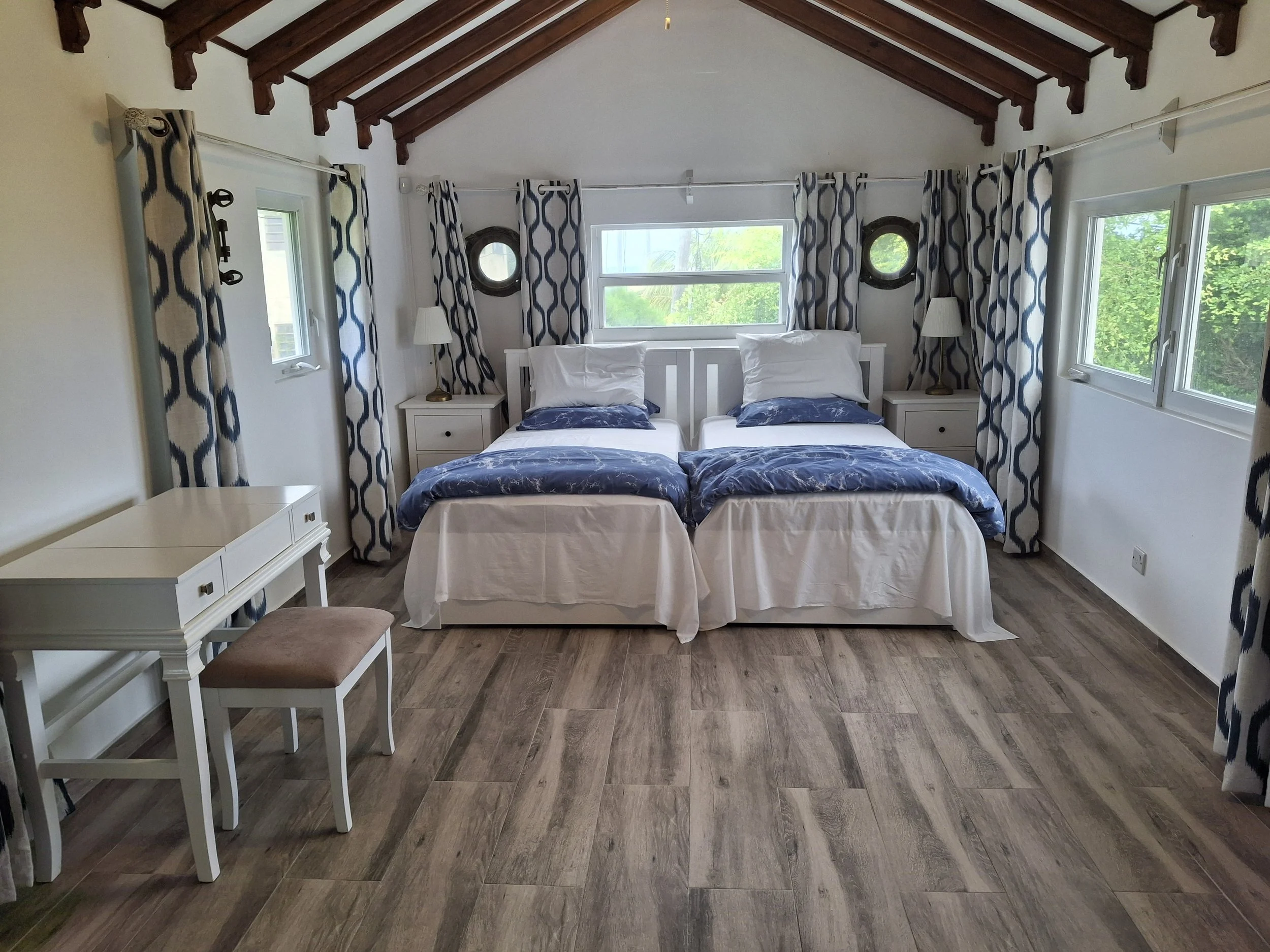 A bedroom with two twin beds pushed together, a white nightstand and lamps on either side. The room has wooden beams on the ceiling, three windows with blue and white patterned curtains, and a small desk with a stool.
