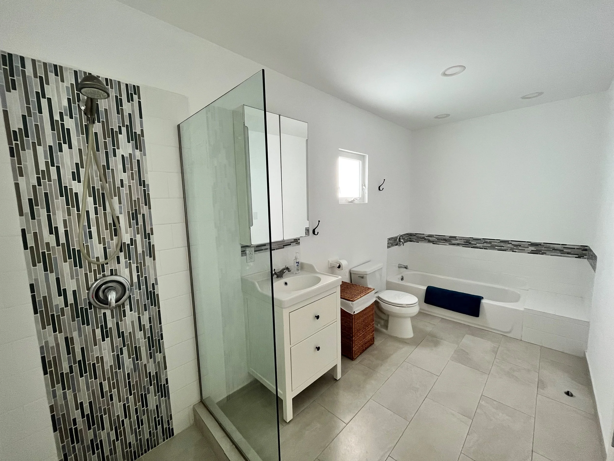 A modern bathroom featuring a walk-in shower with a black and white mosaic tile accent wall, a clear glass shower enclosure, a white vanity with a mirror cabinet, a toilet, and a bathtub with a black towel hanging on the side. The bathroom has white walls, a window, and a light-colored tile floor.