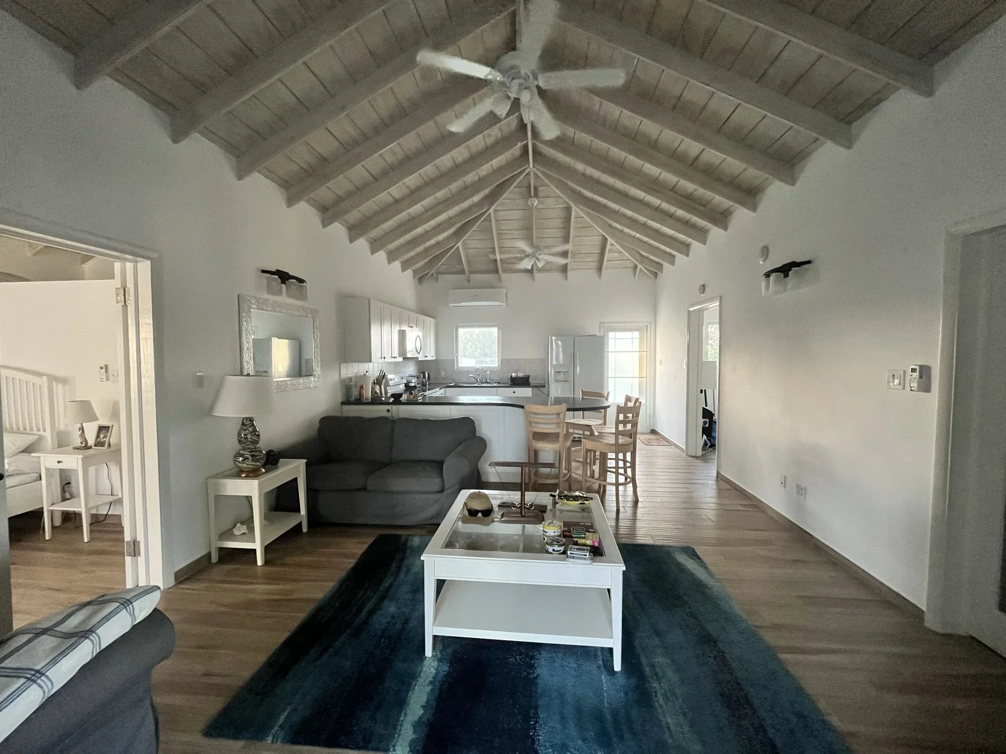 Open-concept living room with vaulted wooden ceiling, gray couch, white coffee table, blue rug, kitchen with white cabinets and bar stools, and bedroom visible through doorway.