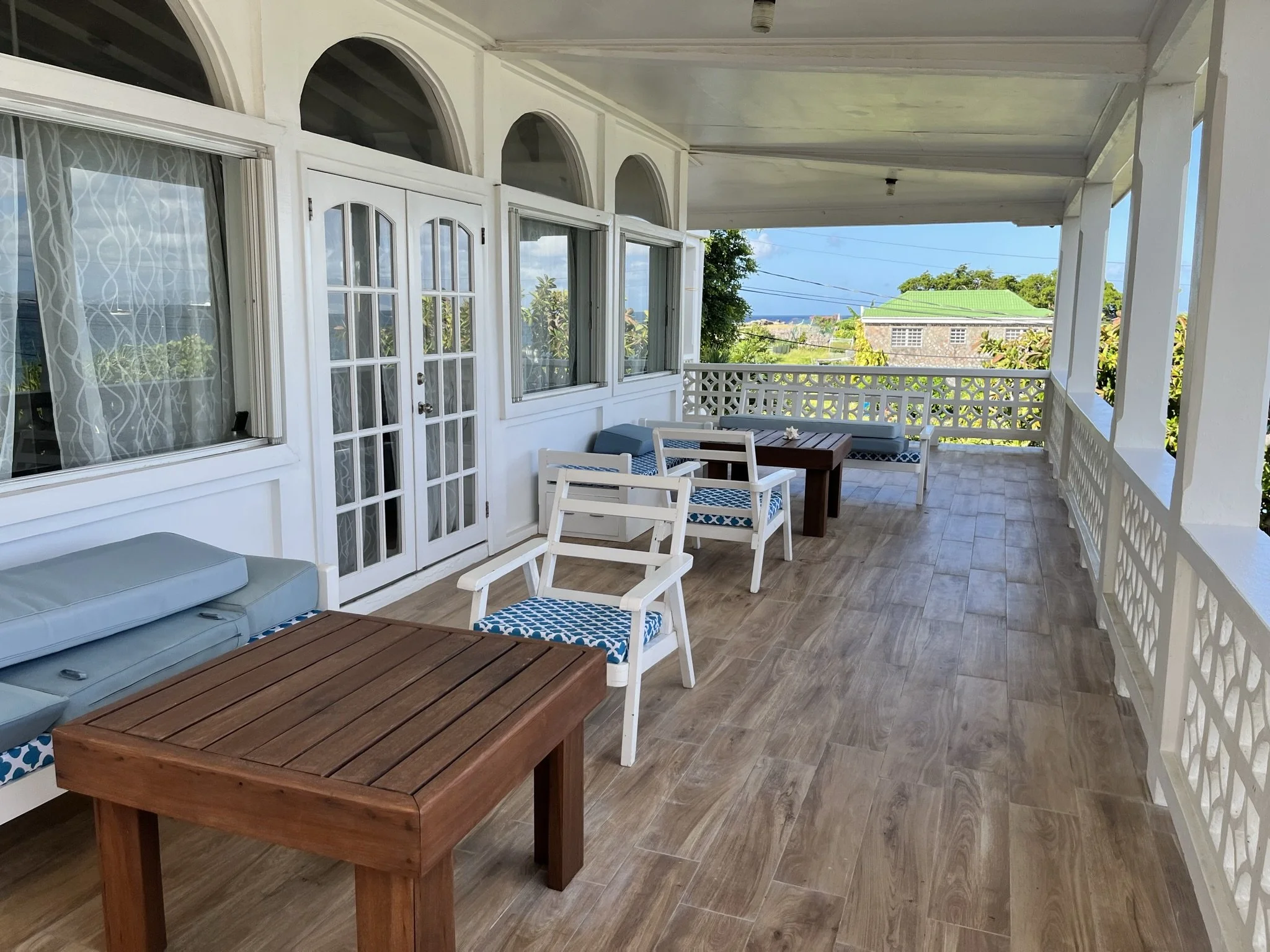 A spacious, covered balcony with wooden tile flooring, white furniture, and great views out to the sea