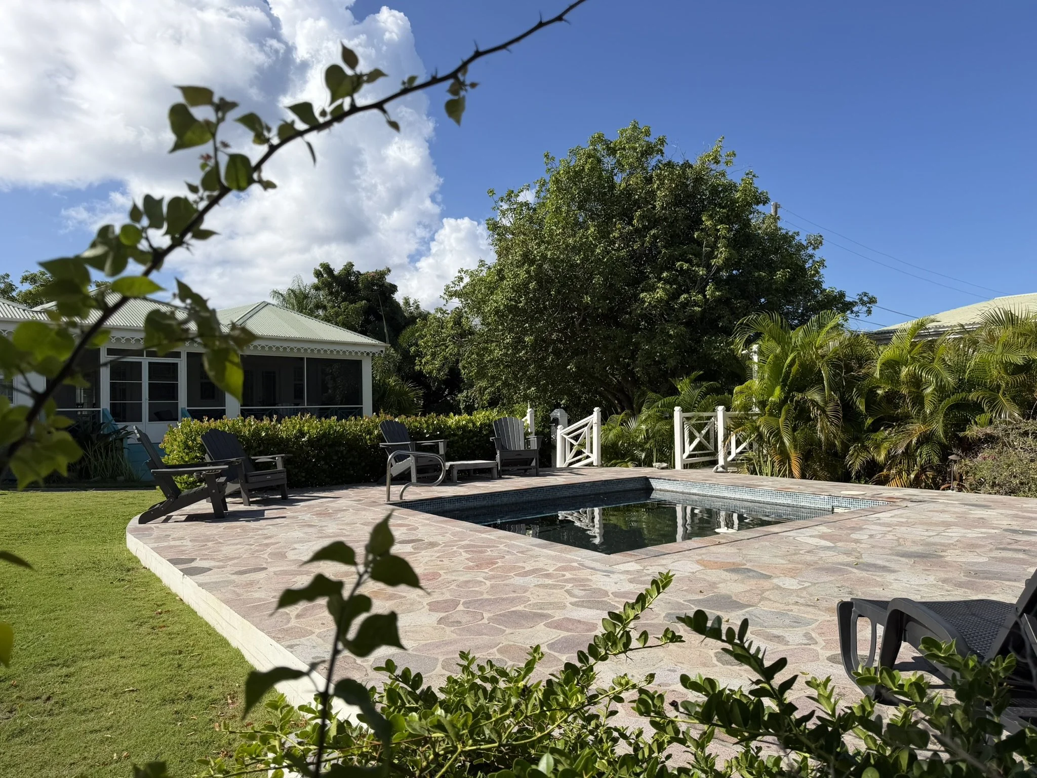 Outdoor pool area with a small rectangular pool, surrounded by a stone patio, black lounge chairs, lush green trees, and a house with a screened porch in the background under a partly cloudy sky.