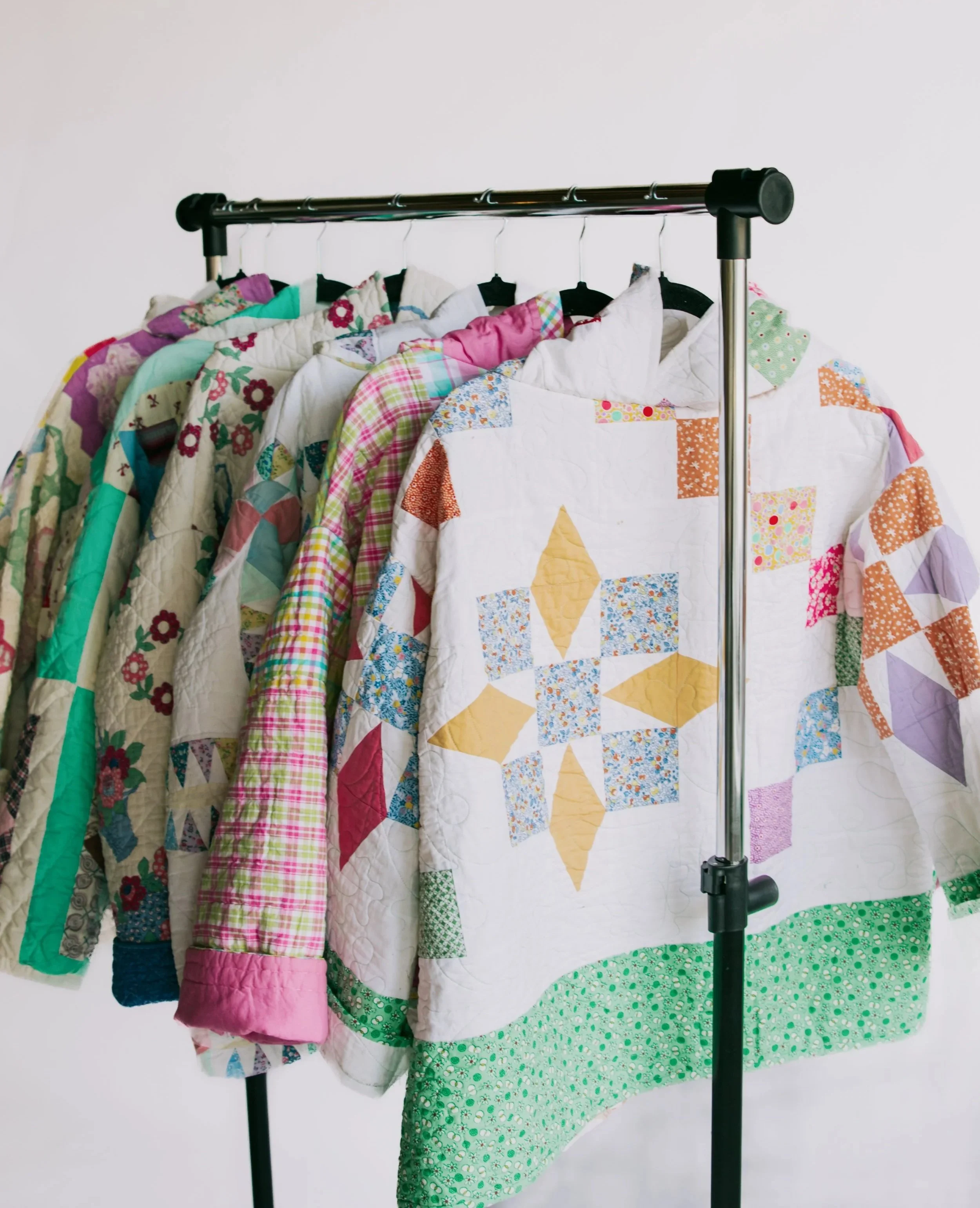 Collection of colorful patchwork quilts hanging on a clothing rack against a plain white background.
