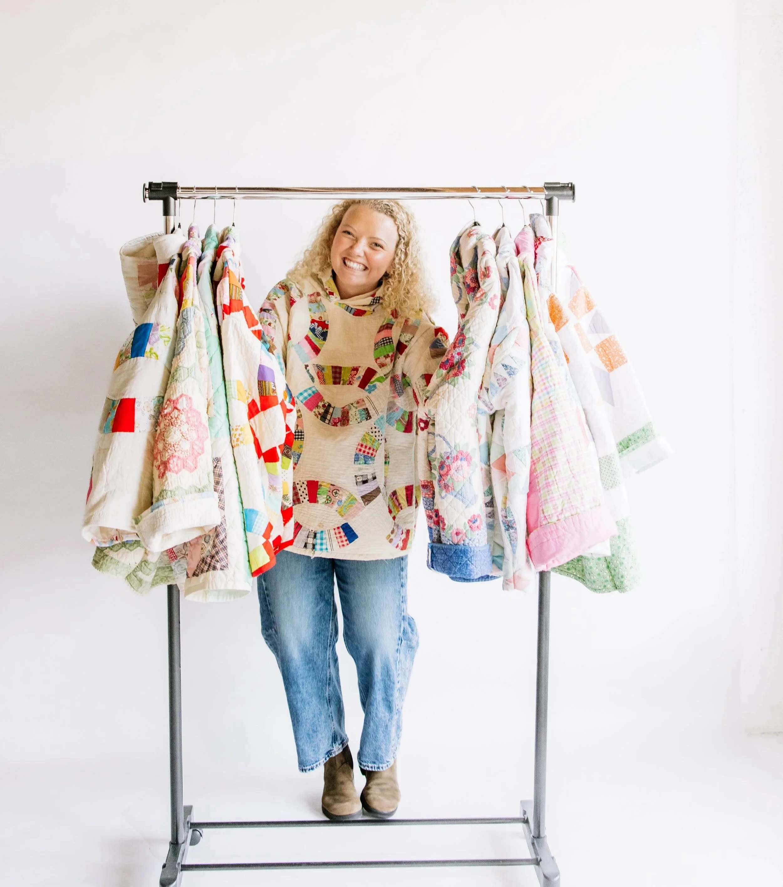 A smiling woman with curly blonde hair stands behind a clothing rack with patchwork quilts hanging on it, against a white background.