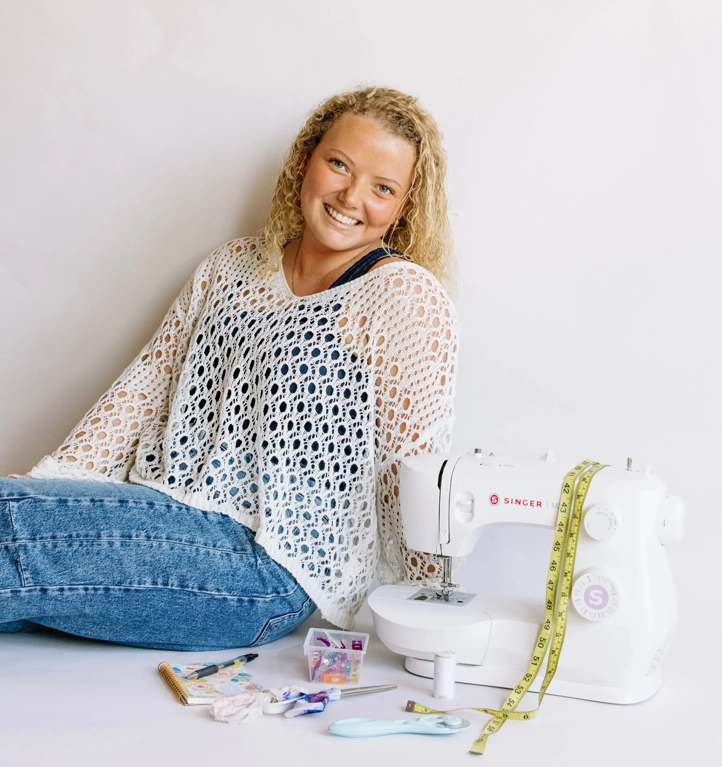 A young woman with curly blonde hair smiling, sitting on the floor, wearing a white crochet top over a navy tank top, and blue jeans, next to a sewing machine with a measuring tape. Sewing supplies are on the floor nearby.