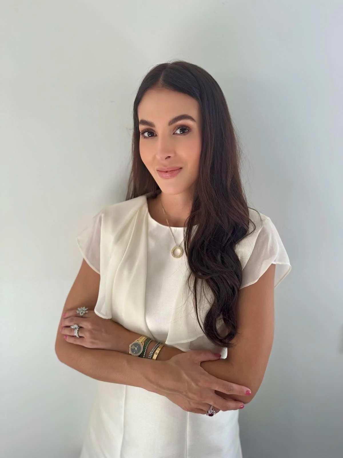 Young woman with long dark wavy hair wearing a white dress and jewelry, standing against a plain background.