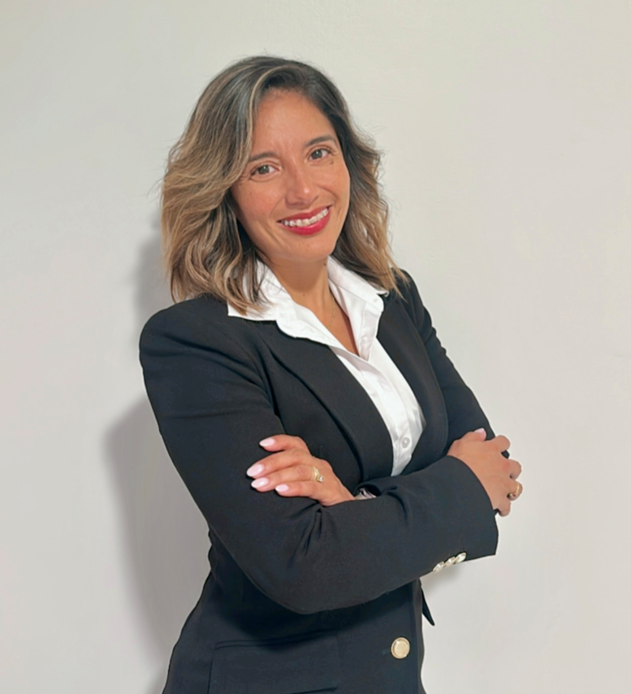 A professional woman with shoulder-length wavy hair, wearing a black blazer and white blouse, smiling with arms crossed against a plain white wall.