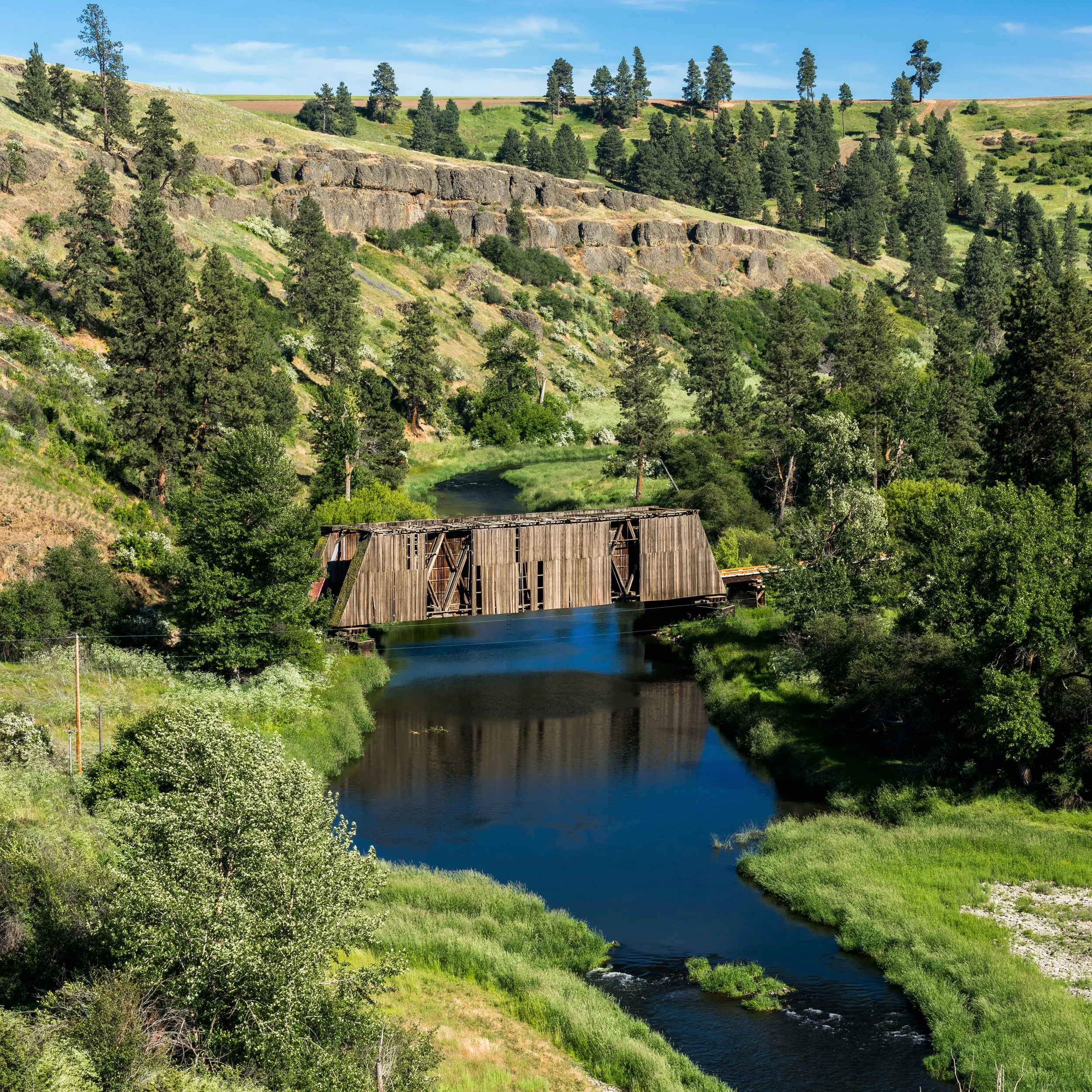 A wooden covered bridge crossing over a river in a lush green valley with trees and hills in the background.