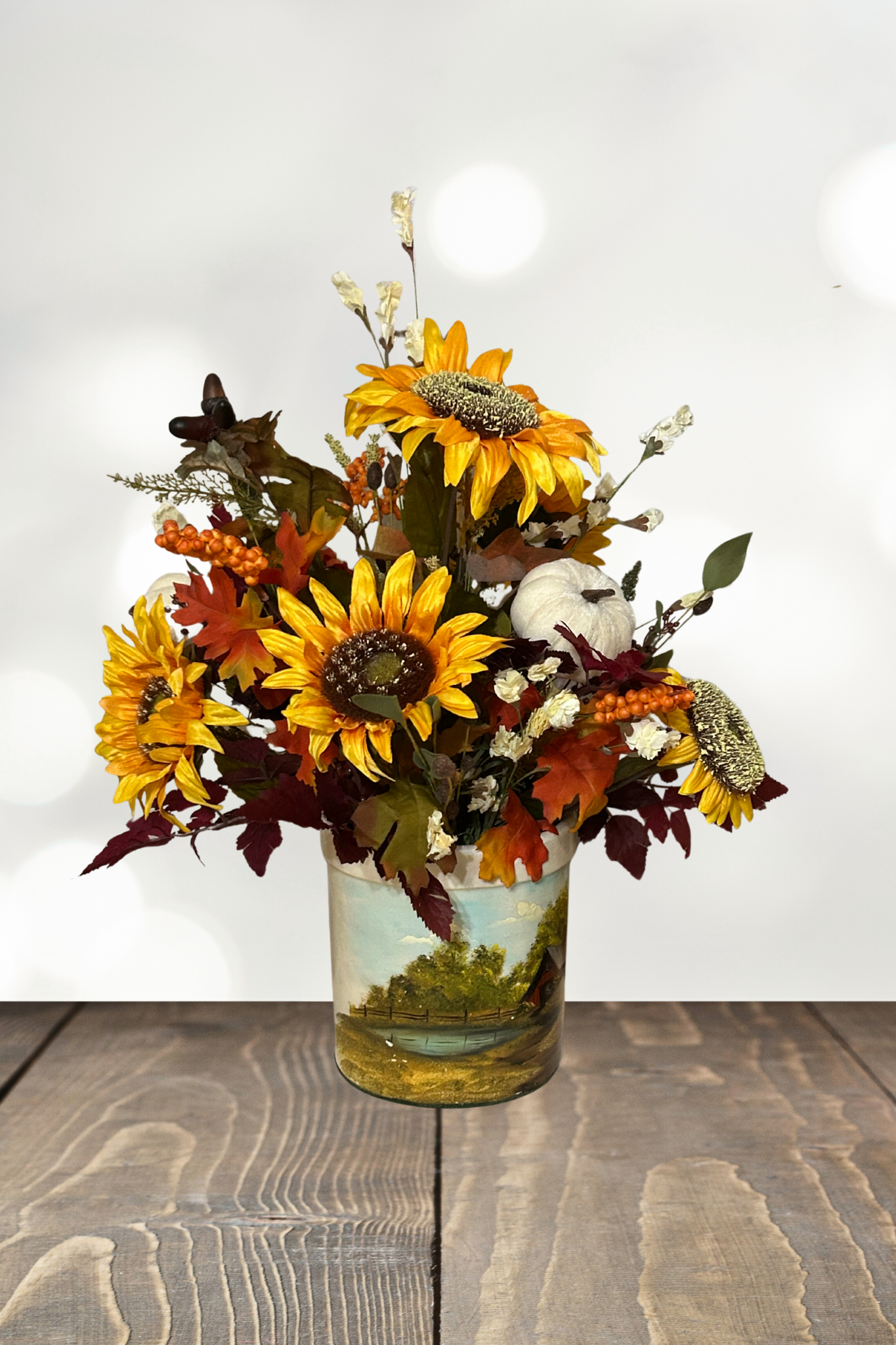 Autumn-themed floral arrangement with sunflowers, white pumpkin, and orange berries in a decorative vase with a landscape scene, on a wooden table.
