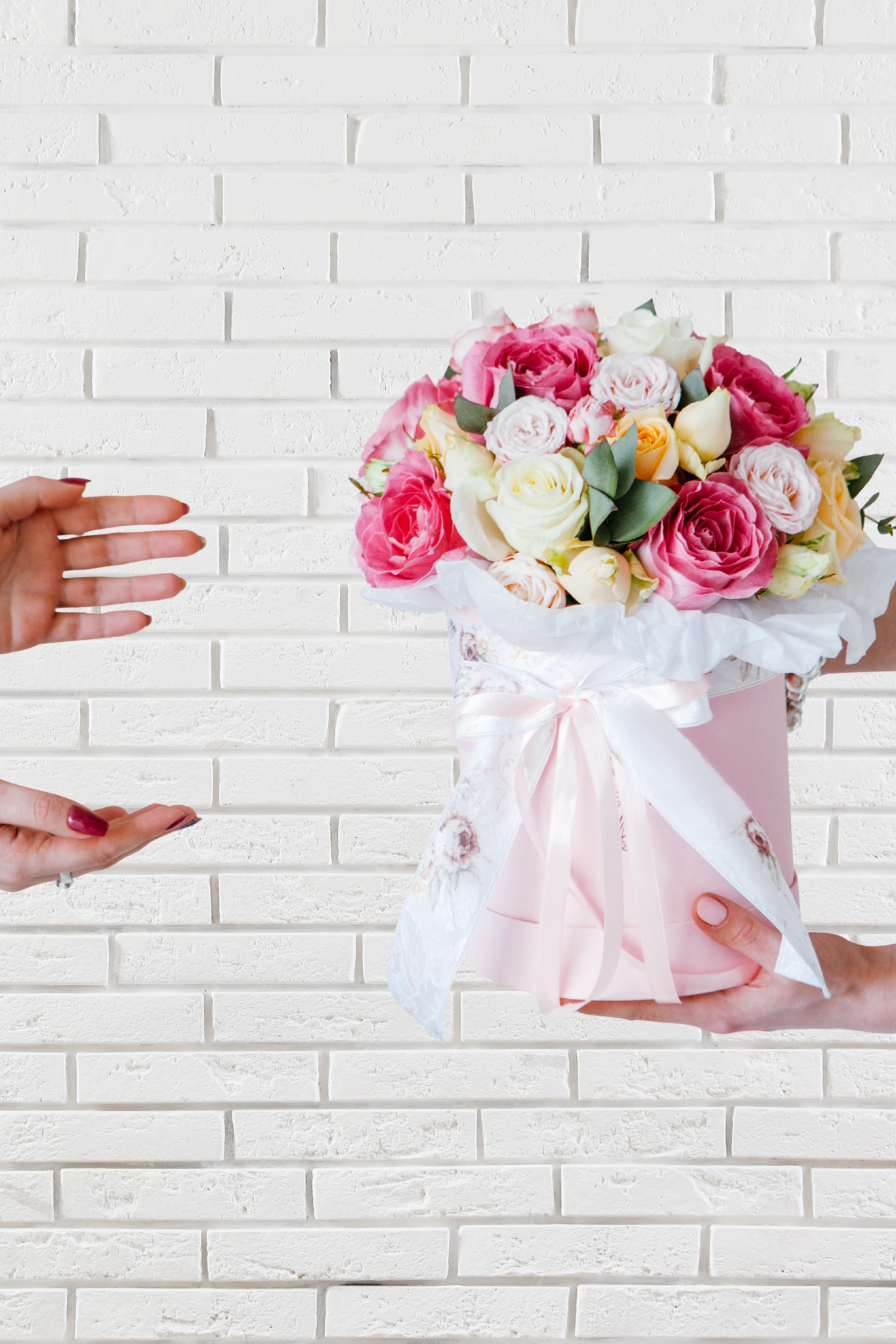 Person handing a pink flower bouquet to another person against a white brick wall.